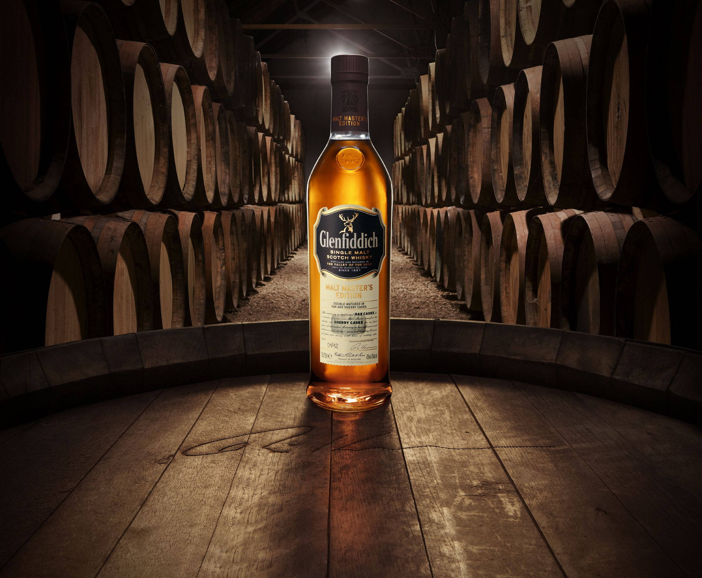 alcohol 桌布 壁紙 — 一瓶發光的 Glenfiddich 威士忌酒瓶，置於木地板上，位於一個光線昏暗、排列著陳年橡木桶的倉庫中央。