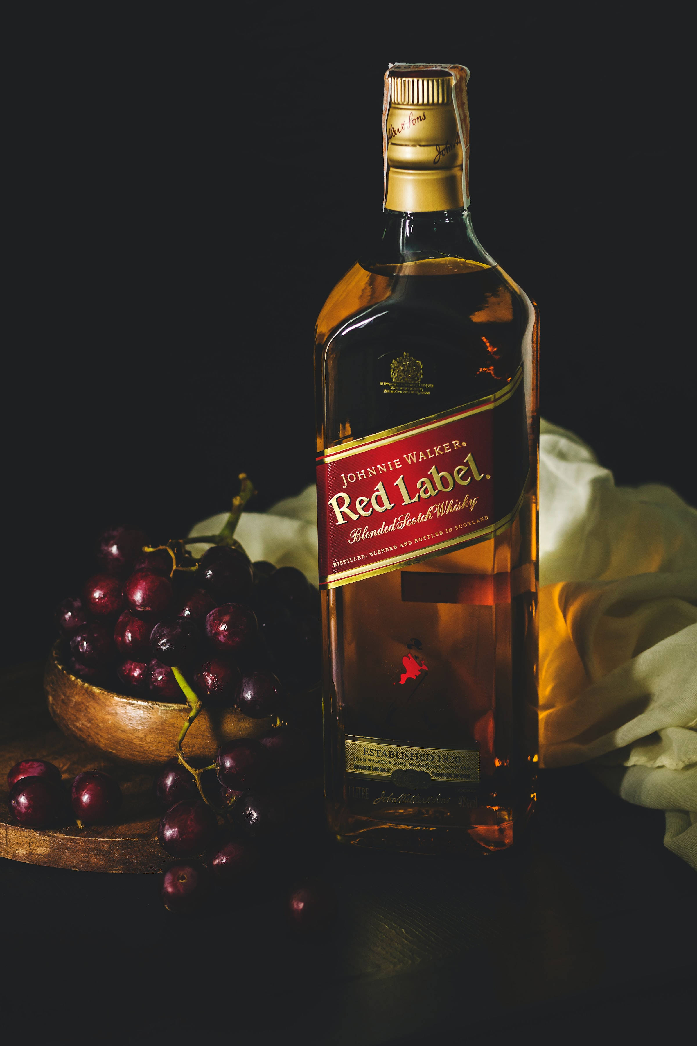 alcohol 桌布 壁紙 — 一瓶約翰走路紅牌威士忌在黑色背景下，溫暖地發光，旁邊放著一個裝有黑葡萄的木碗。