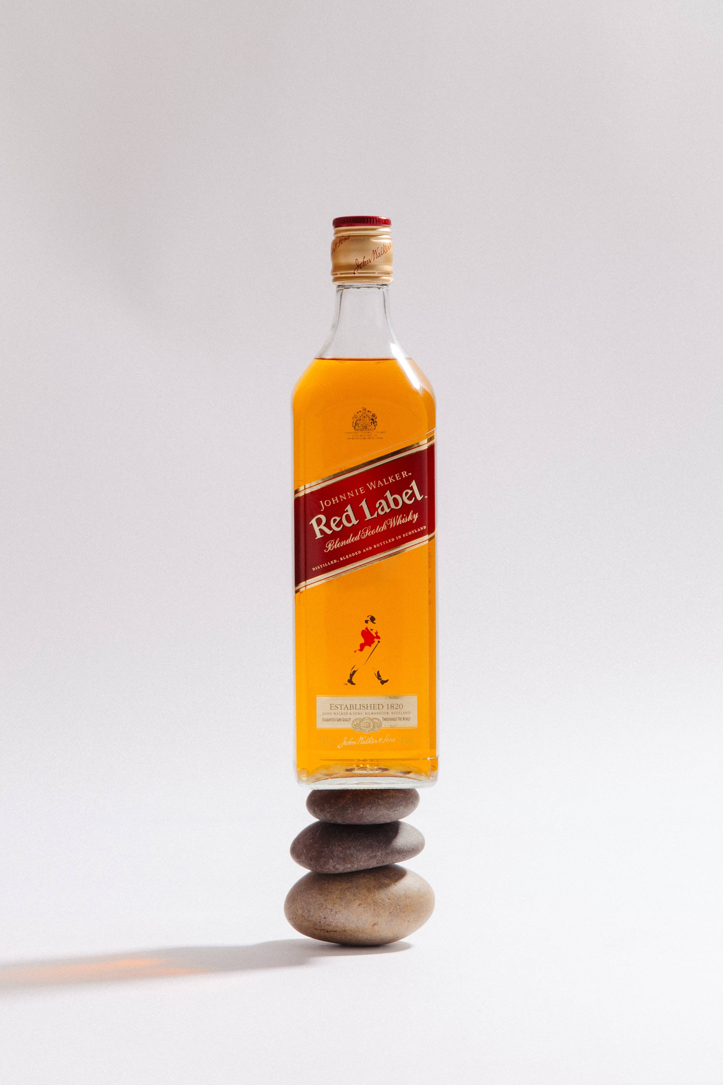 alcohol 桌布 壁紙 — 一瓶約翰走路紅牌威士忌酒瓶平衡地放在一堆光滑石頭上，背景為純白色。