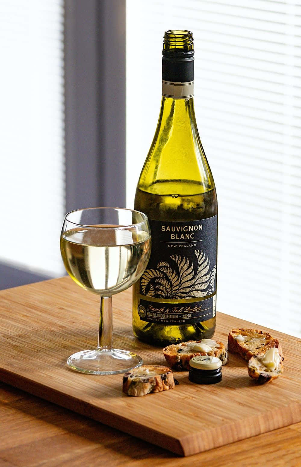White Wine 桌布 壁紙 — 一瓶紐西蘭白蘇維濃葡萄酒和裝滿酒的酒杯放在木製砧板上，旁邊有塗了奶油的克羅斯蒂尼，靠近百葉窗。