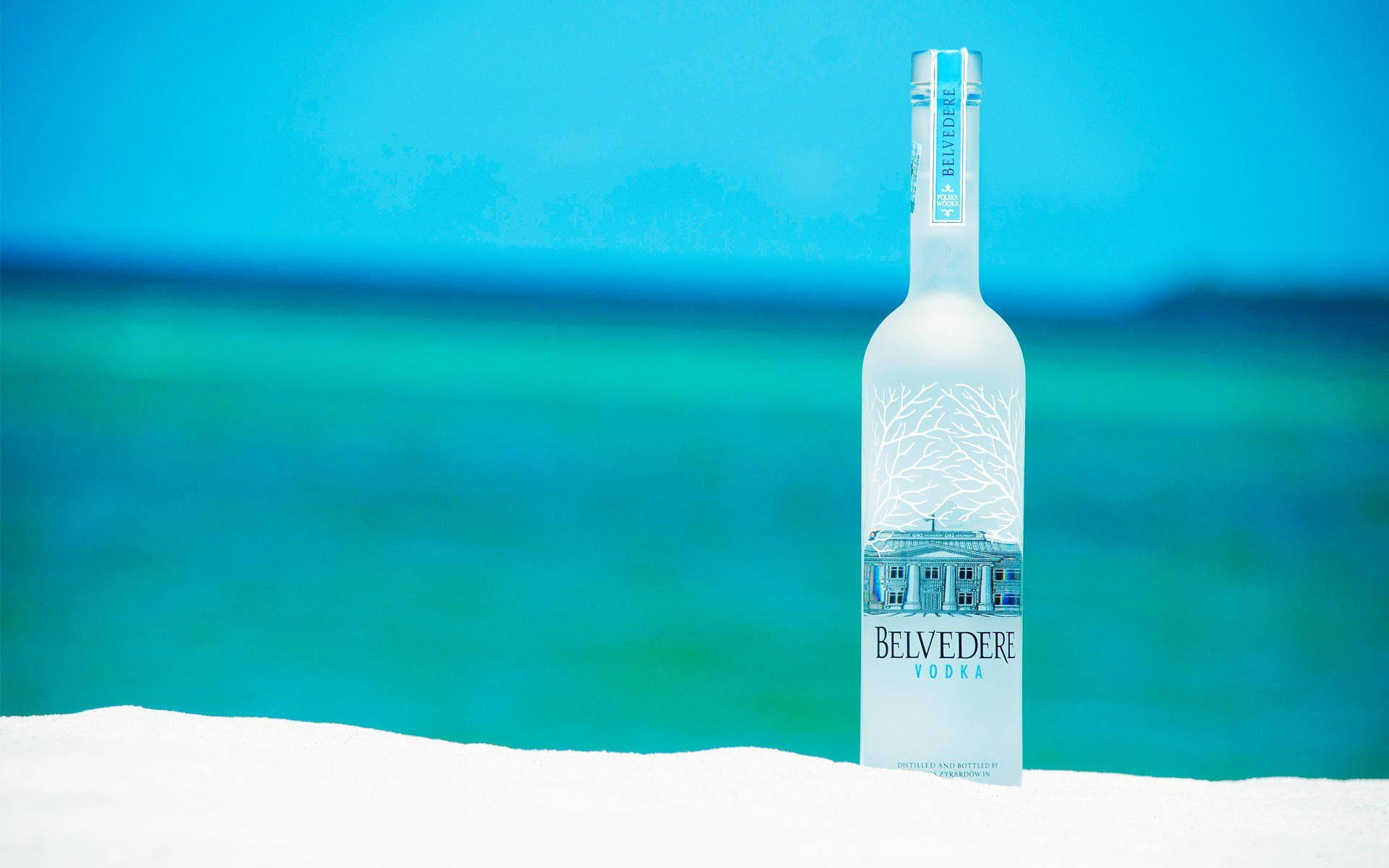 alcohol 桌布 壁紙 — 一瓶結霜的Belvedere伏特加酒瓶立在白色沙灘上，背景是充滿活力的藍綠色海洋和清澈的藍天。