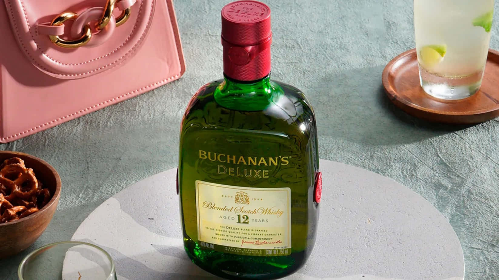 alcohol 桌布 壁紙 — 一瓶綠色Buchanan's Deluxe威士忌放在中央，旁邊有一個粉紅色手提包、萊姆雞尾酒和椒鹽脆餅，放在有紋理的灰色表面上。