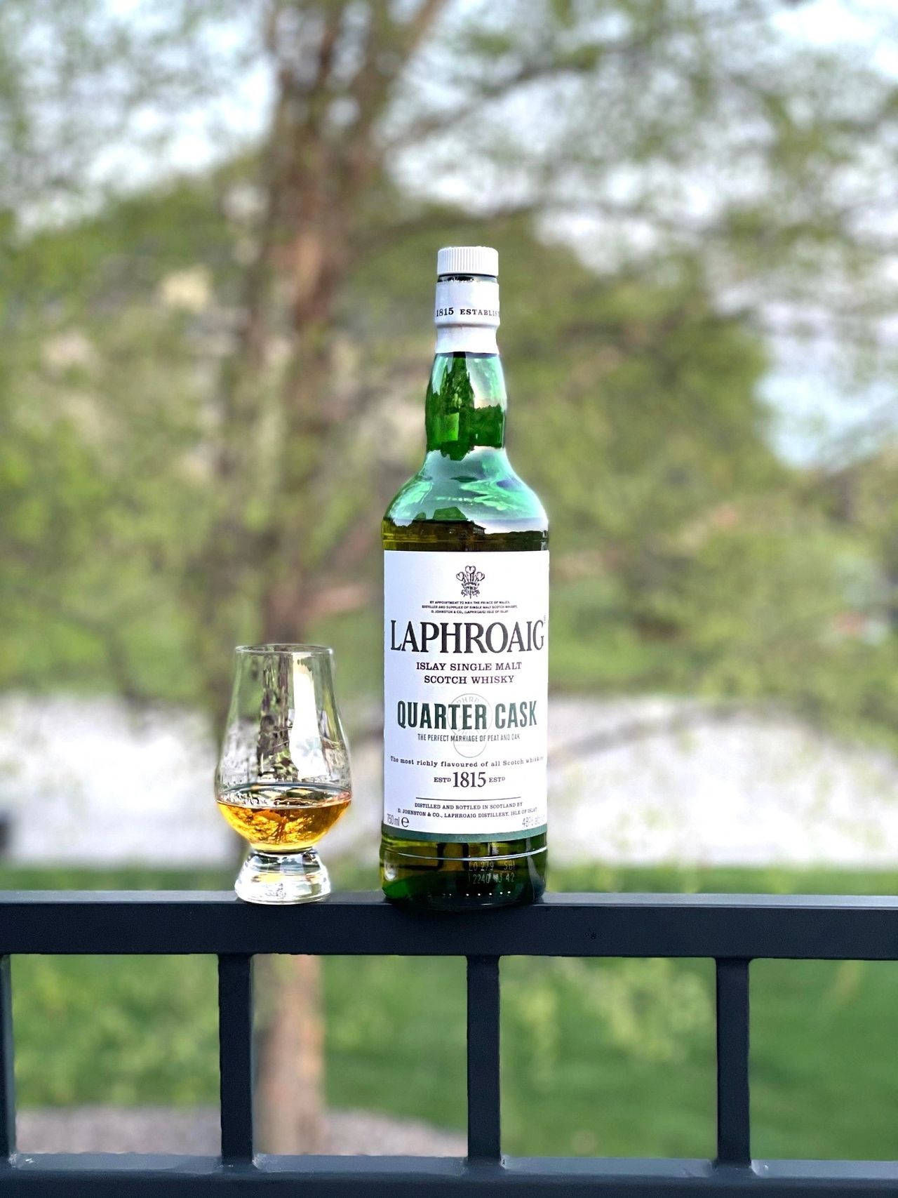 手機 桌布 壁紙 — 一瓶綠色Laphroaig Quarter Cask威士忌和一個裝滿的酒杯放在黑色欄杆上，背景是模糊的綠色樹木和水面。