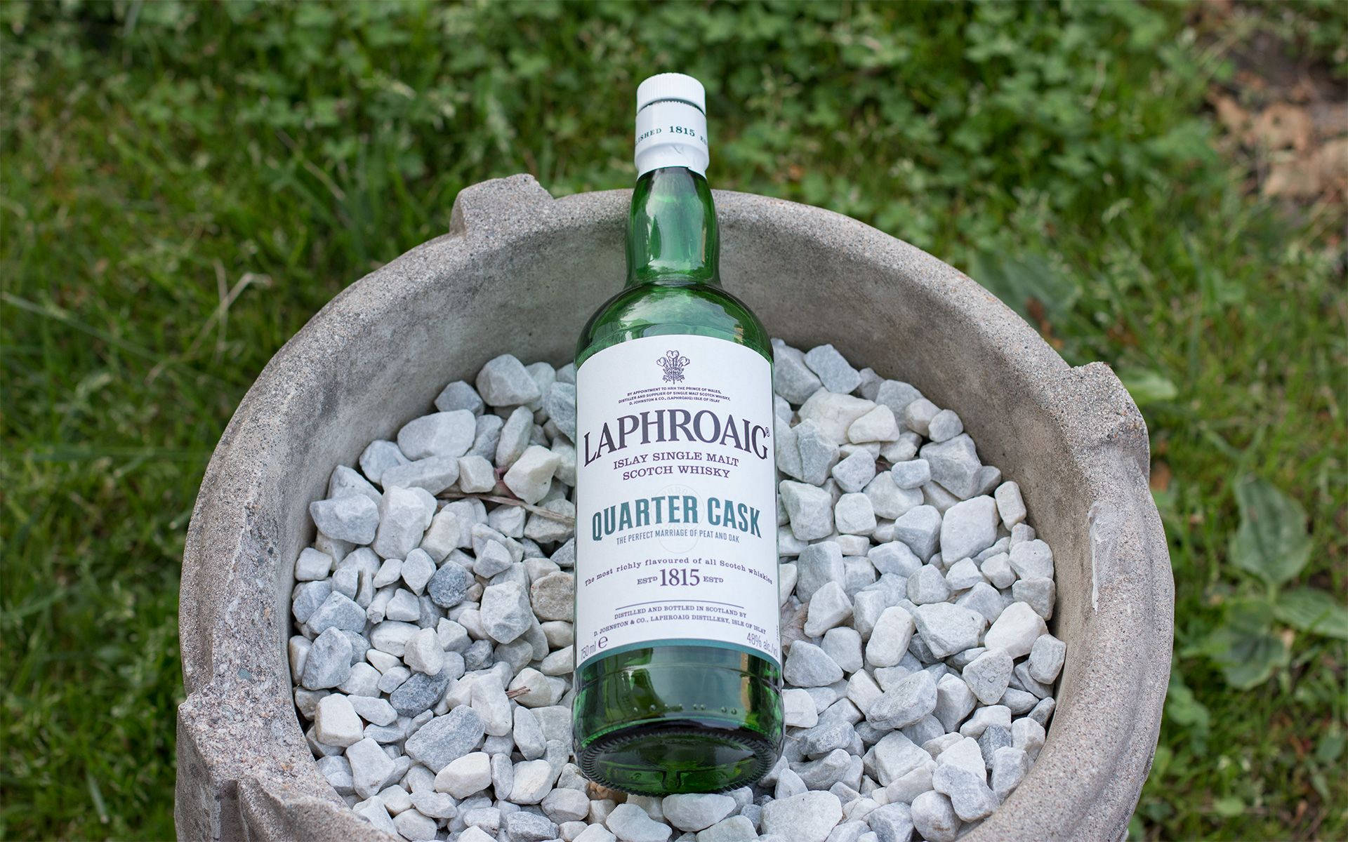 iPhone 桌布 壁紙 — 一瓶綠色Laphroaig Quarter Cask威士忌放在一個裝滿灰色鵝卵石的石製花盆裡，置於草地上。