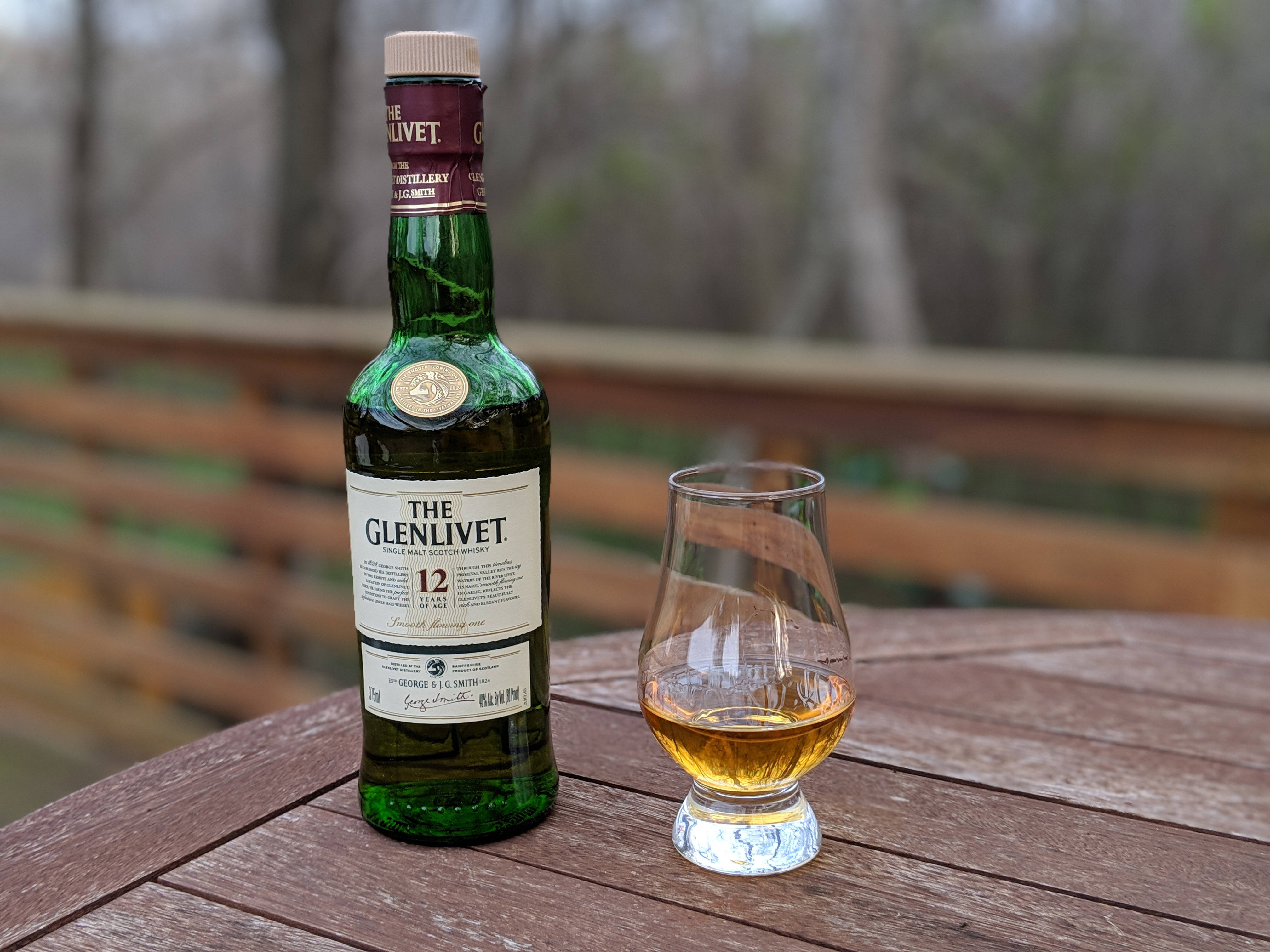 glass 桌布 壁紙 — 一瓶綠色The Glenlivet 12 Year Old威士忌和一杯琥珀色液體放在戶外風化的木桌上。