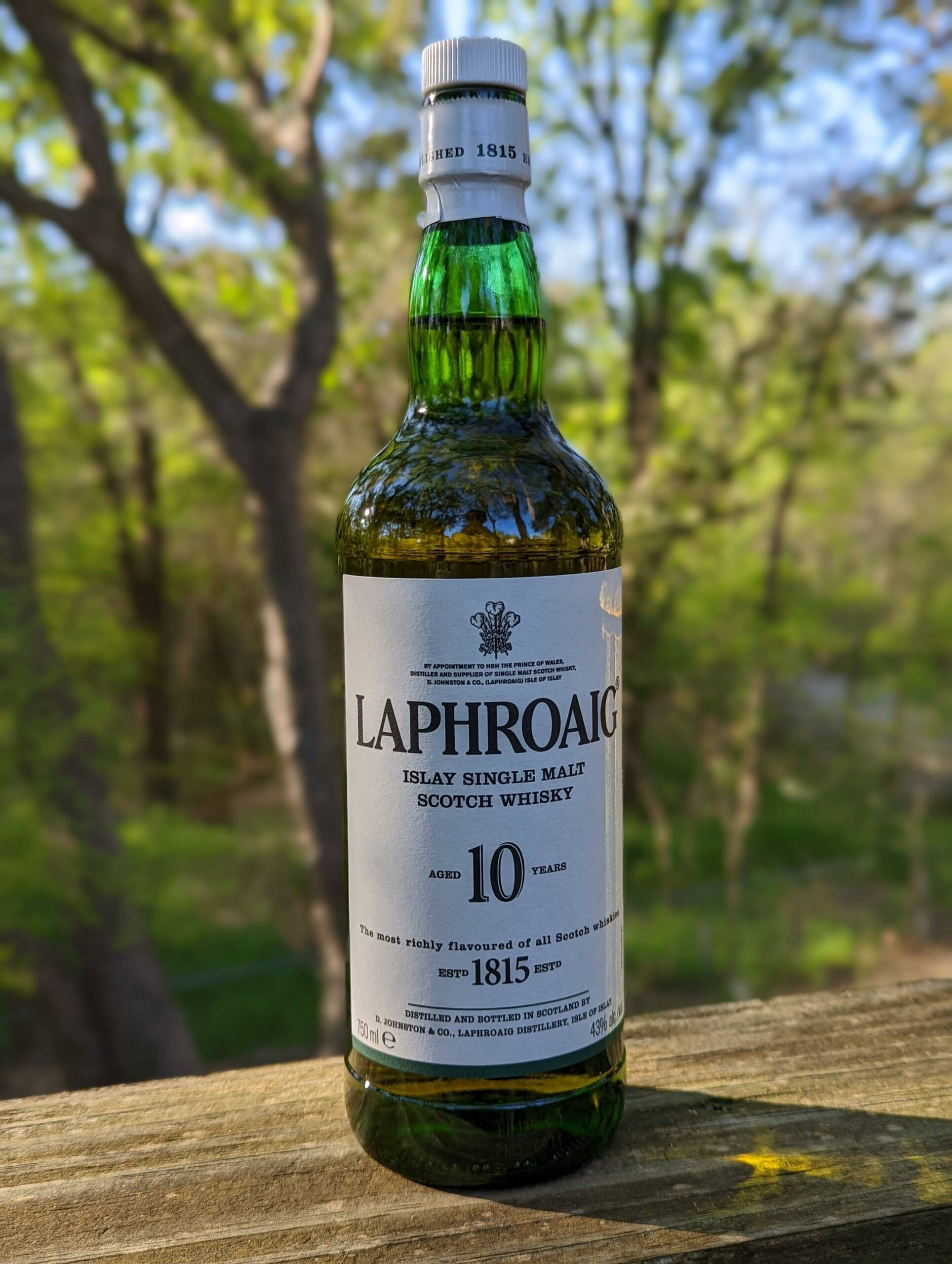 電腦 桌布 壁紙 — 一瓶綠色玻璃瓶裝的Laphroaig 10年艾雷島單一麥芽蘇格蘭威士忌放在戶外的木桌上，背景有模糊的樹木和陽光。