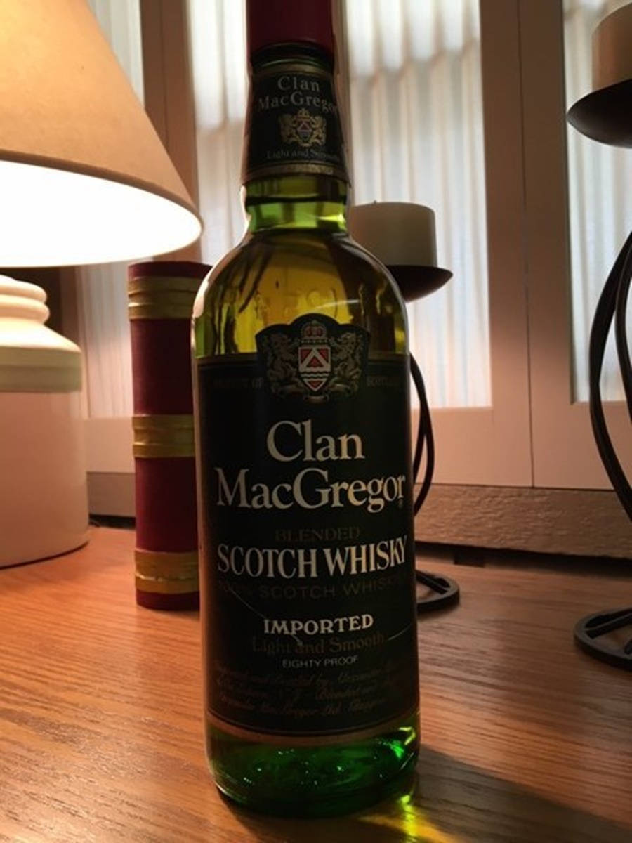 Whiskey 桌布 壁紙 — 一瓶綠色的Clan MacGregor調和蘇格蘭威士忌放在木桌上，旁邊有一盞點亮的燈和一本書。