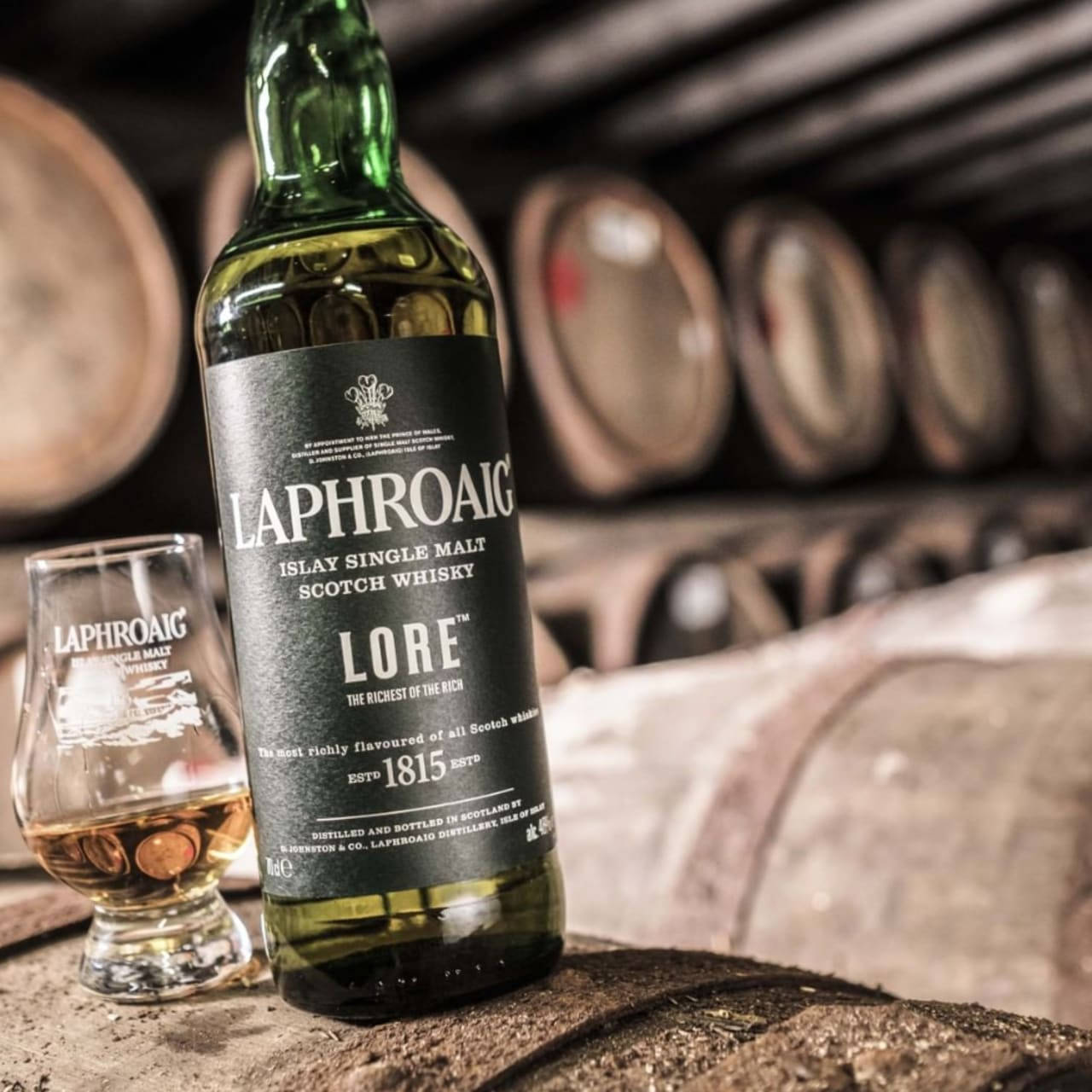 電腦 桌布 壁紙 — 一瓶綠色的Laphroaig Lore威士忌放在一個木桶上，旁邊有一個品酒杯，背景是一個燈光昏暗的蒸餾廠，有許多橡木桶排列在後面。