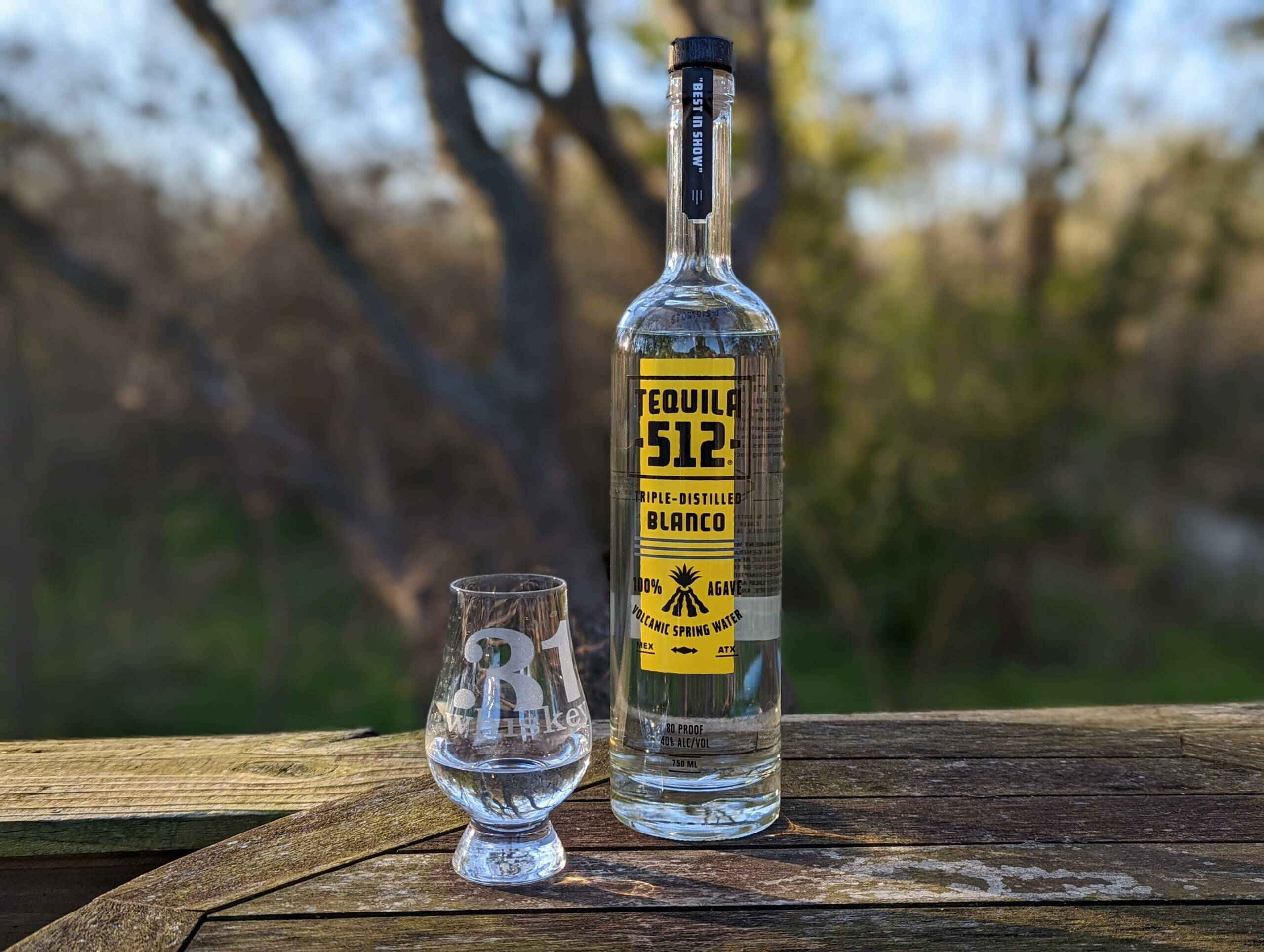 drinks 桌布 壁紙 — 一瓶透明的 512 Tequila Blanco 和一個品牌品酒杯放在戶外一張風化的木桌上，背景是模糊的樹木。
