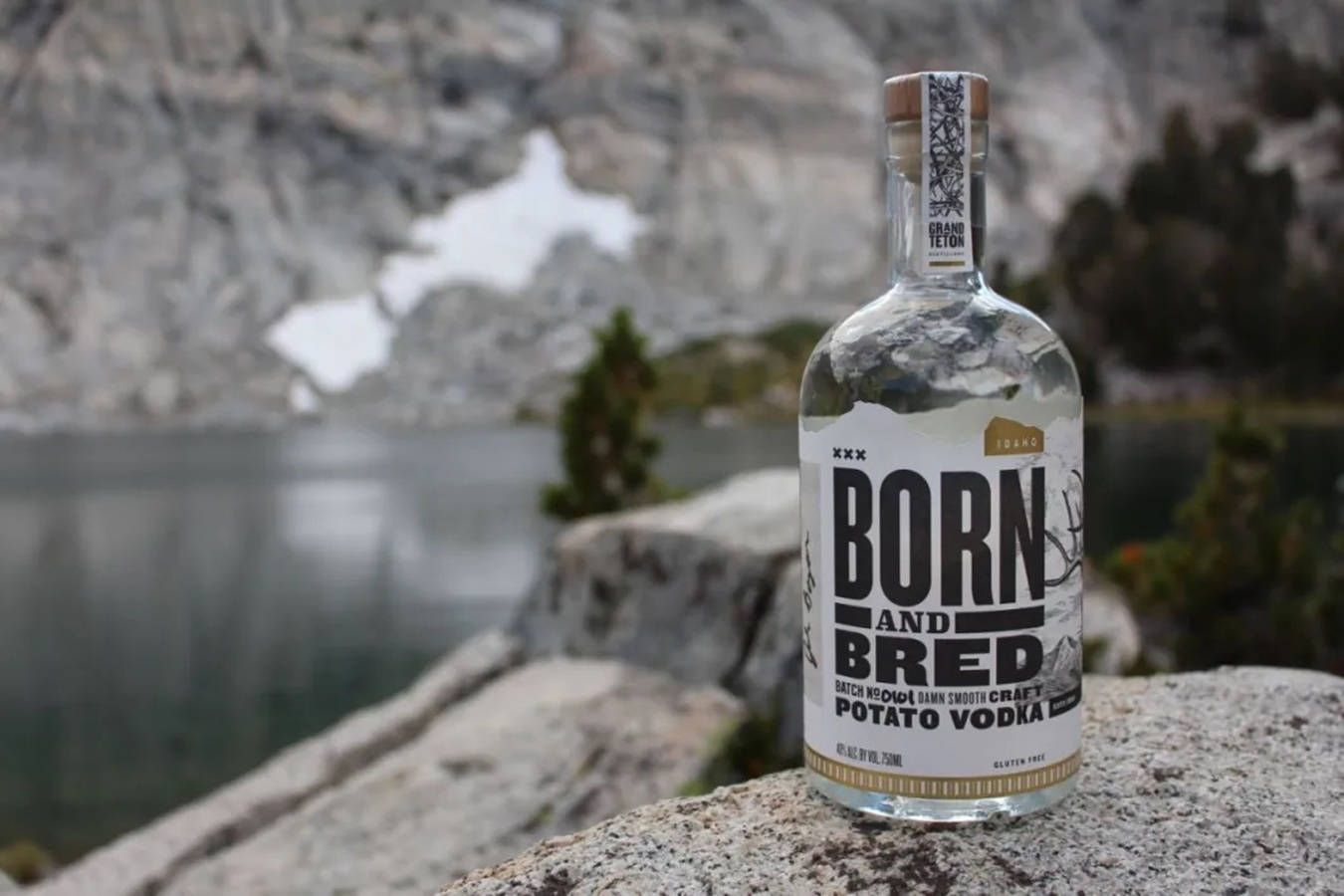 Lake 桌布 壁紙 — 一瓶透明的 Born and Bred 馬鈴薯伏特加酒放在岩石架上，俯瞰著寧靜的山湖，背景有雪斑。