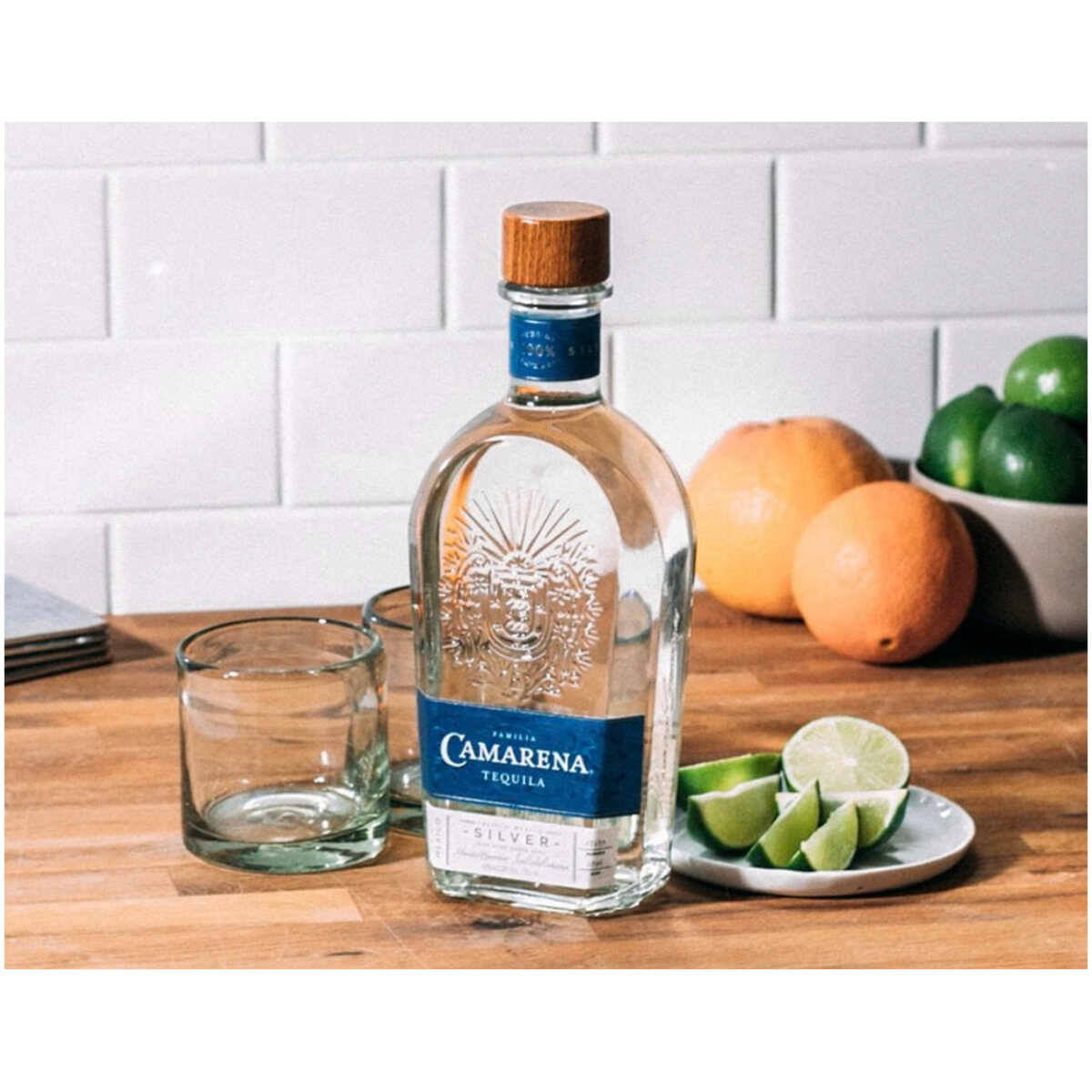 Tequila 桌布 壁紙 — 一瓶透明的 Camarena Silver 龍舌蘭酒放在木質檯面上，旁邊有萊姆角和柑橘類水果，背景是白色瓷磚牆。