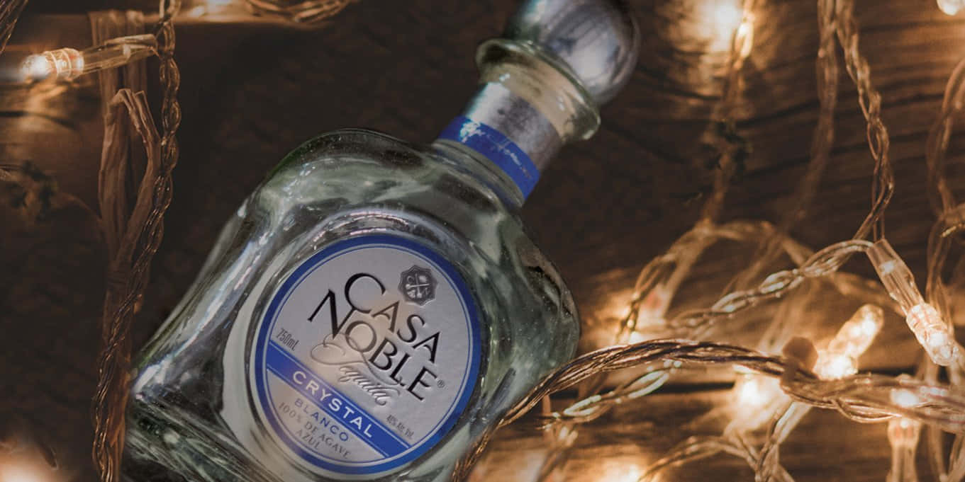 Tequila 桌布 壁紙 — 一瓶透明的 Casa Noble Crystal Blanco 龍舌蘭酒放在木質表面上，周圍環繞著溫暖發光的串燈。