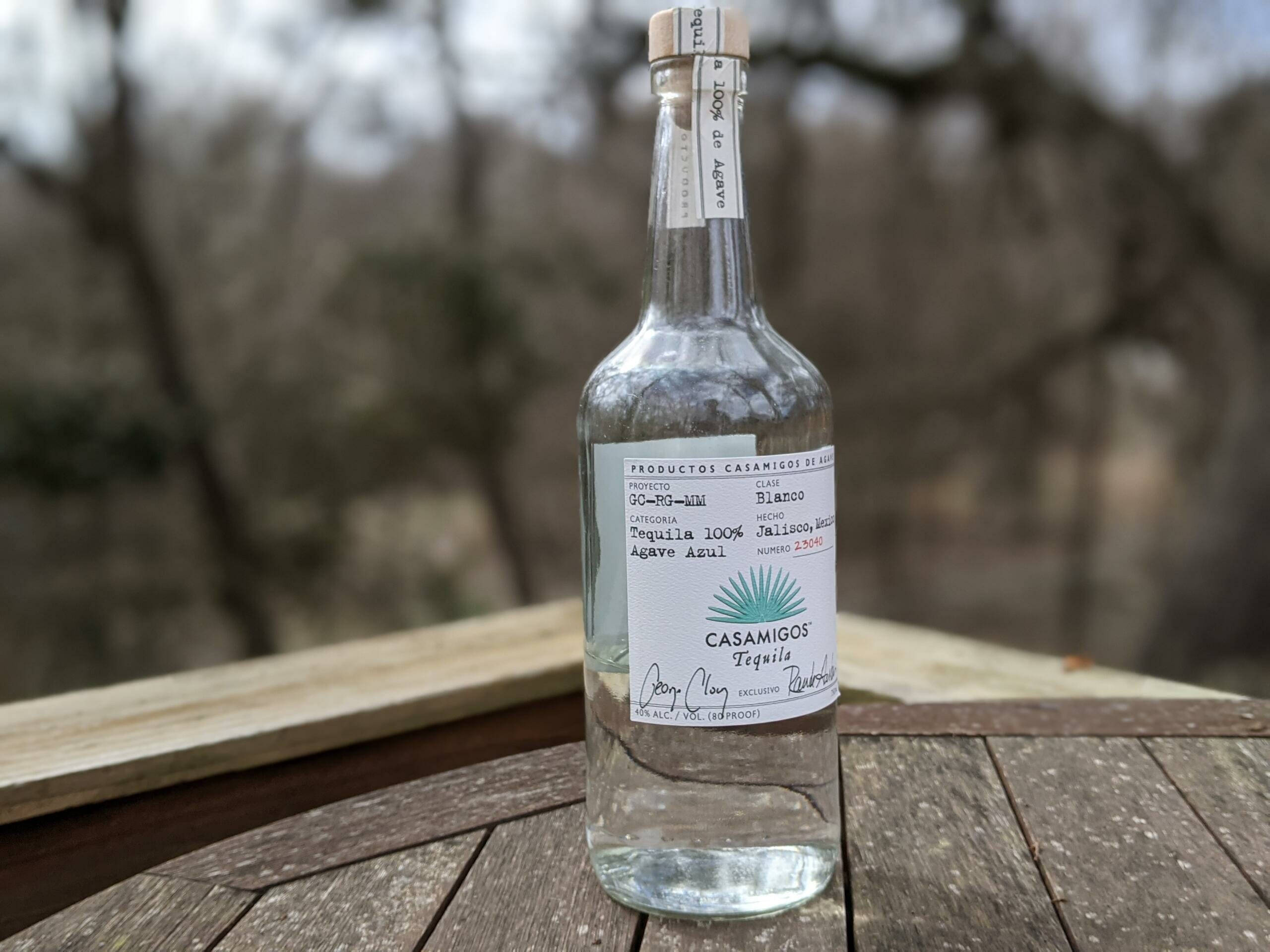 bottle 桌布 壁紙 — 一瓶透明的 Casamigos Blanco 龍舌蘭酒放在一張風化的木桌上，背景是模糊的樹木。