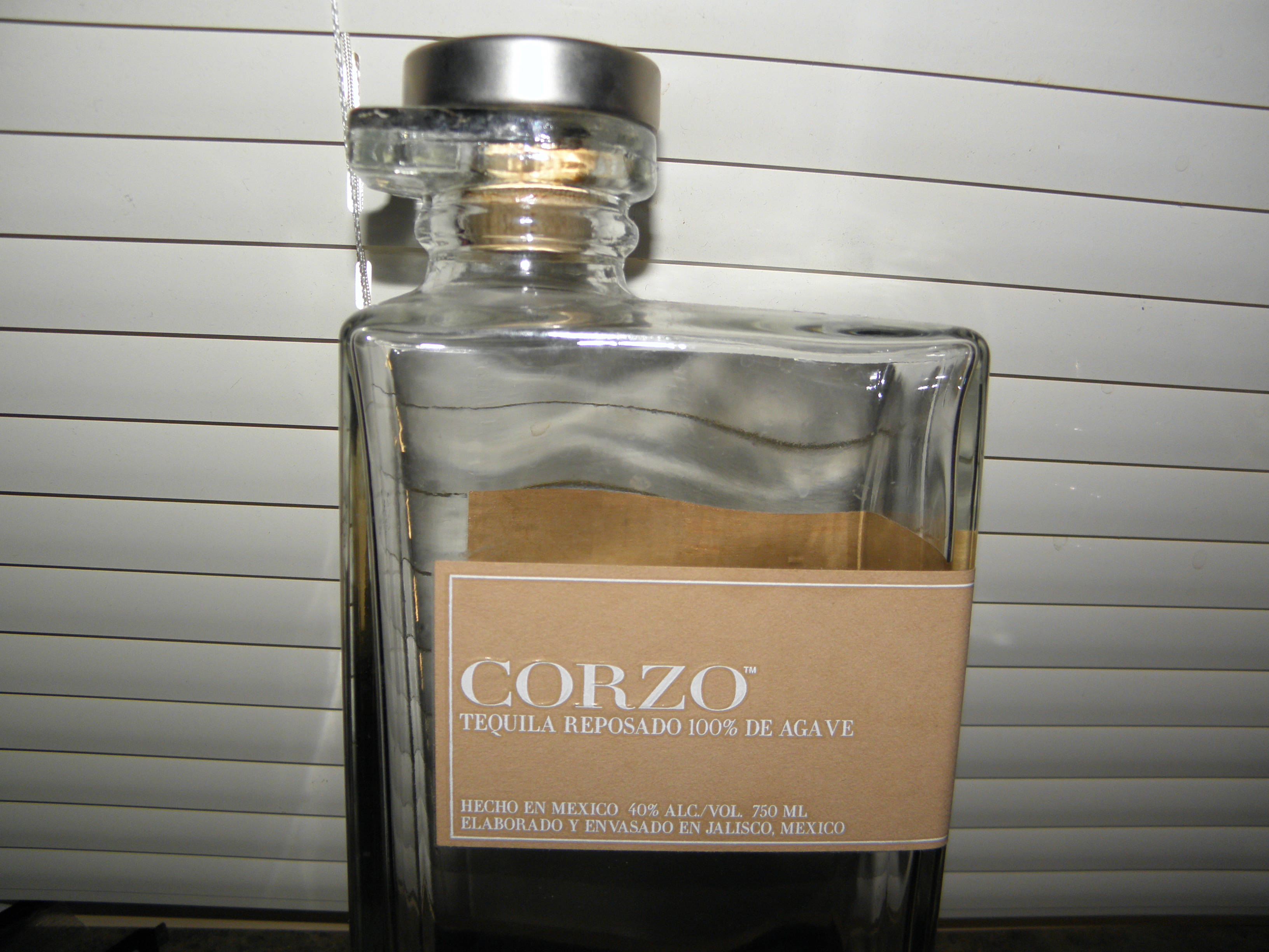 bottle 桌布 壁紙 — 一瓶透明的 Corzo Tequila Reposado 玻璃瓶，帶有棕色紙標籤，放在白色橫向百葉窗簾前。
