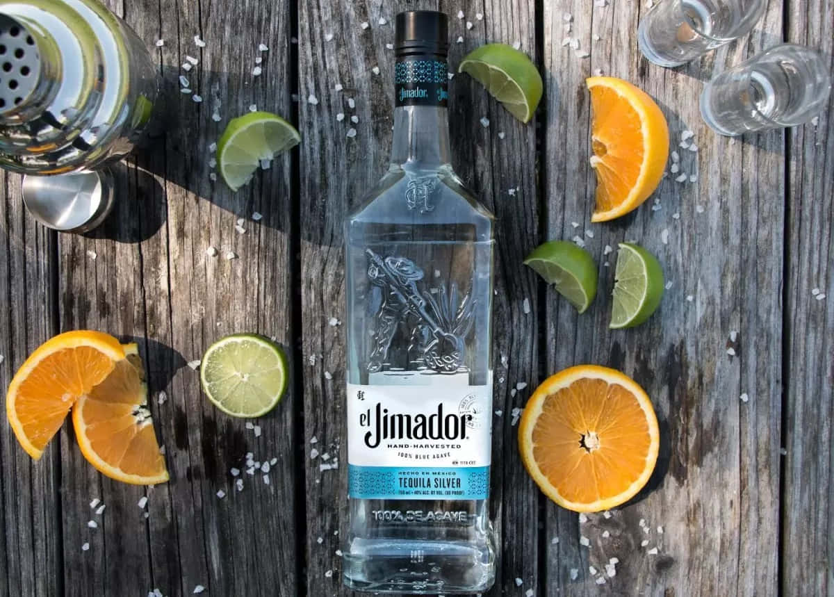drinks 桌布 壁紙 — 一瓶透明的 El Jimador 龍舌蘭酒放在一張風化的木桌中央，周圍環繞著柑橘片和鹽。