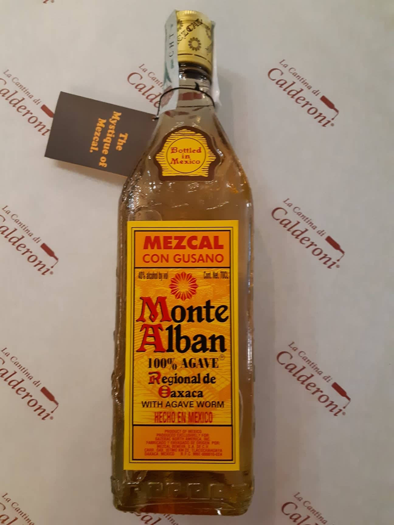 Drink 桌布 壁紙 — 一瓶透明的 Monte Alban 梅斯卡爾酒，內有龍舌蘭蟲，放在有品牌標誌的紙背景上。