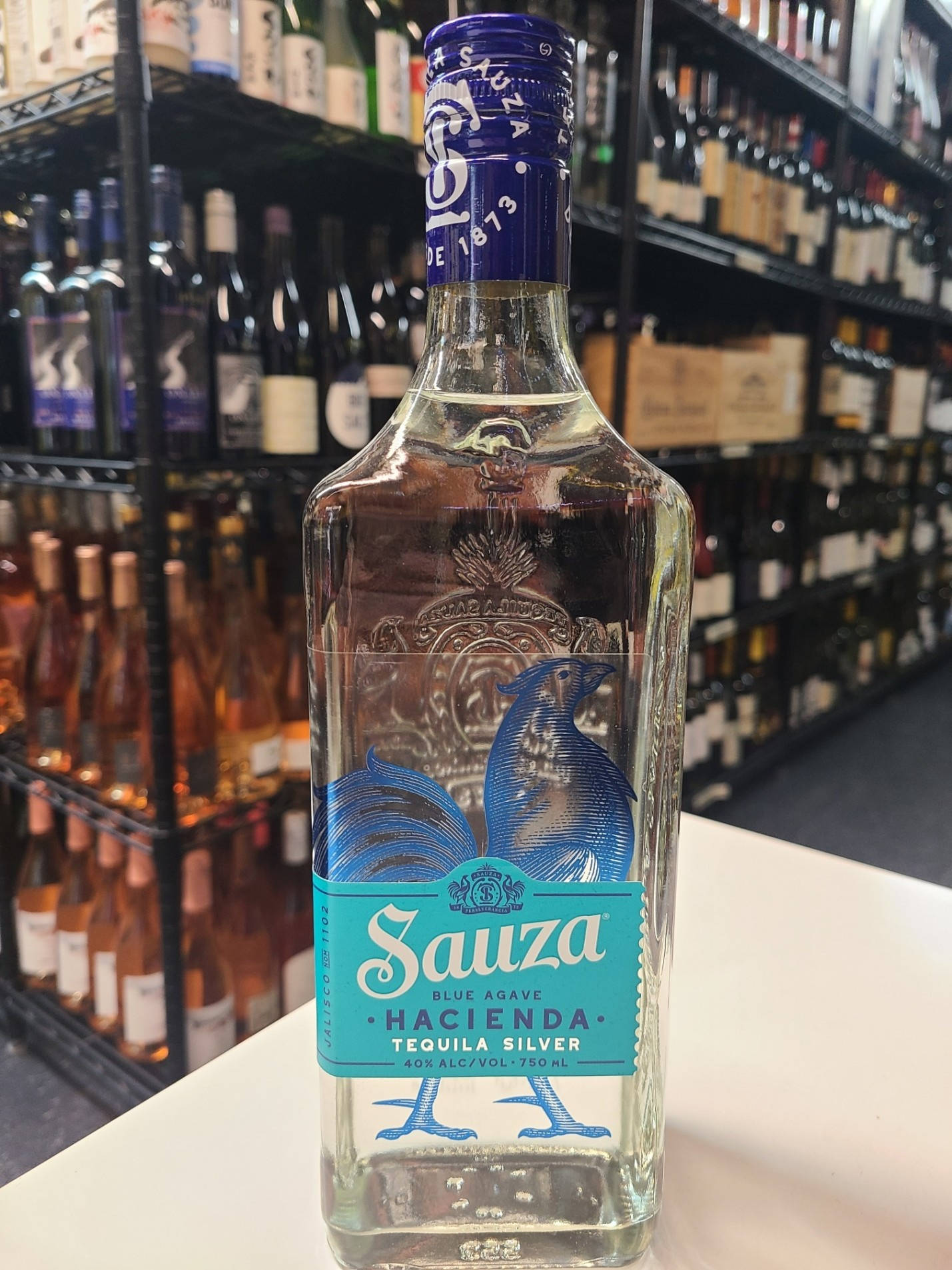 高清 桌布 壁紙 — 一瓶透明的 Sauza Hacienda Blue Agave Tequila Silver 放在白色櫃檯上，背景是擺滿酒瓶的貨架。