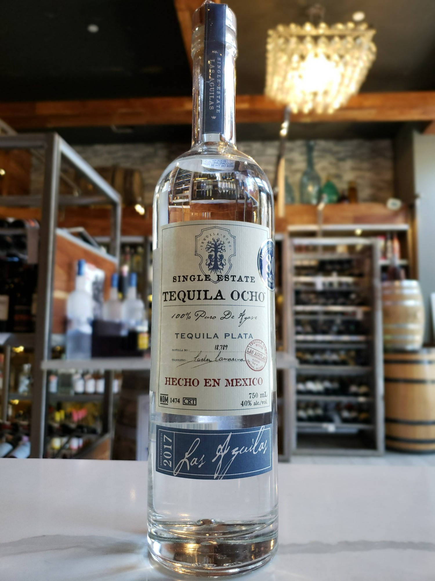 bottle 桌布 壁紙 — 一瓶透明的 Single Estate Tequila Ocho 放在白色檯面上，位於一間燈光溫暖、有木樑和吊燈的酒吧中央。