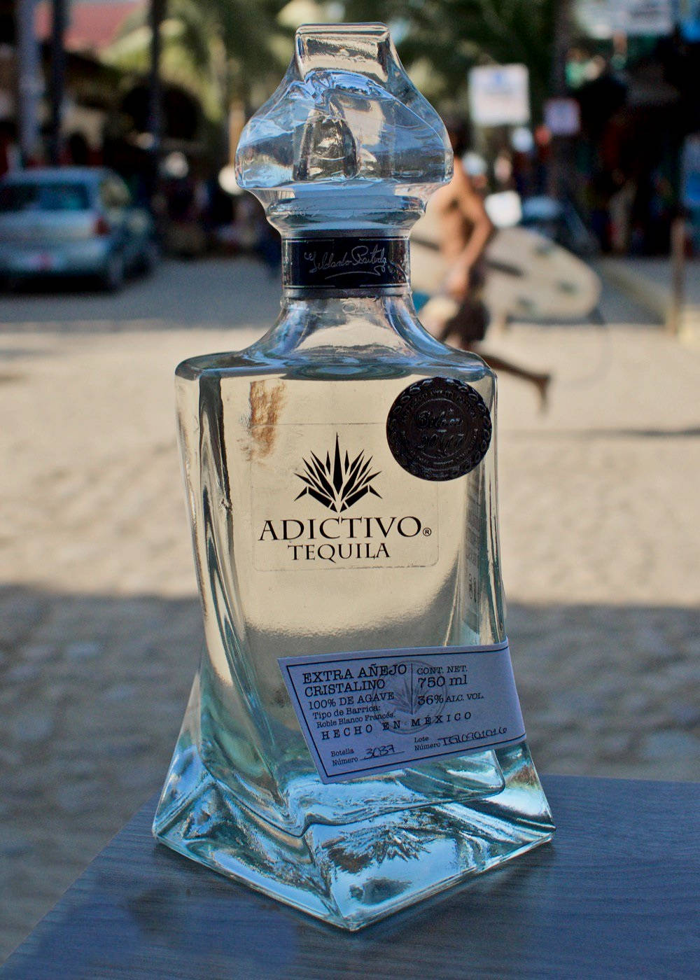 iPhone 桌布 壁紙 — 一瓶透明的Adictivo Tequila玻璃瓶放在戶外的桌子上，背景是模糊的海濱街道場景。