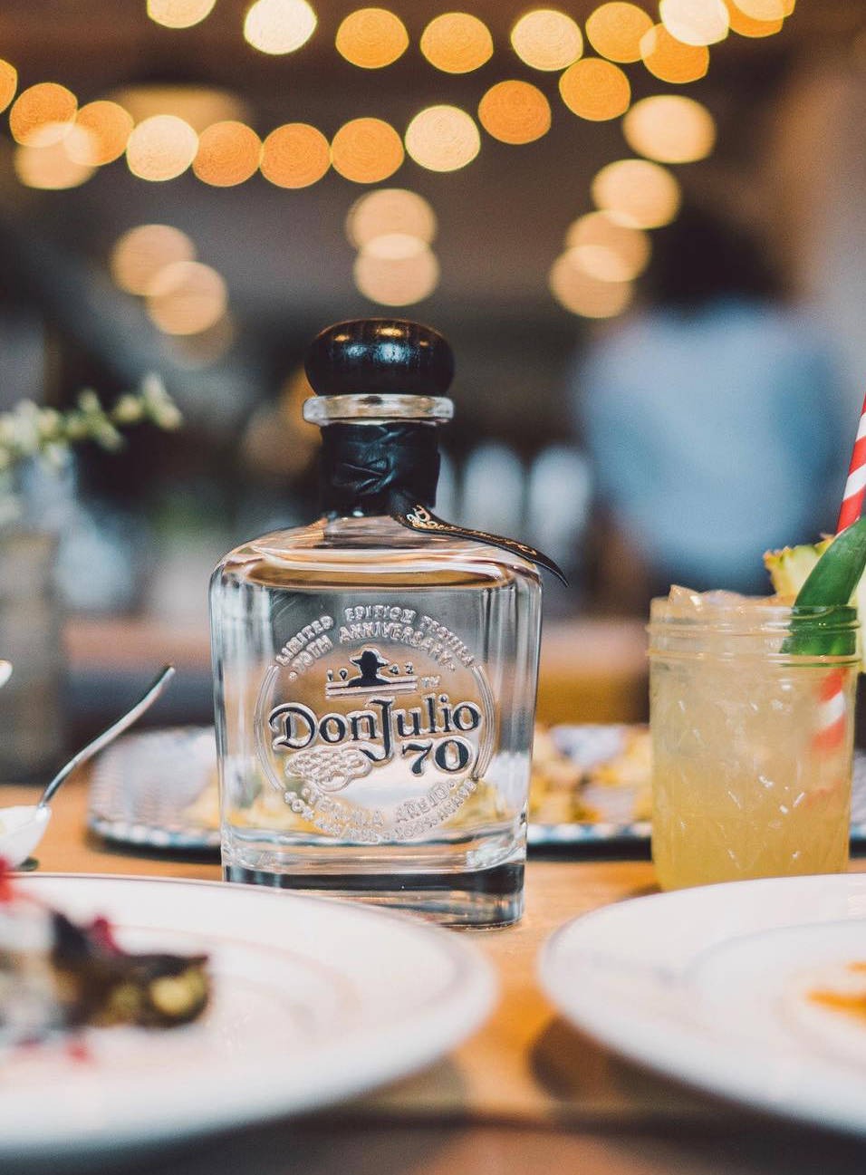 Drink 桌布 壁紙 — 一瓶透明的Don Julio 70龍舌蘭酒放在木桌上，旁邊有熱帶雞尾酒和甜點，在溫暖的散景燈光下。