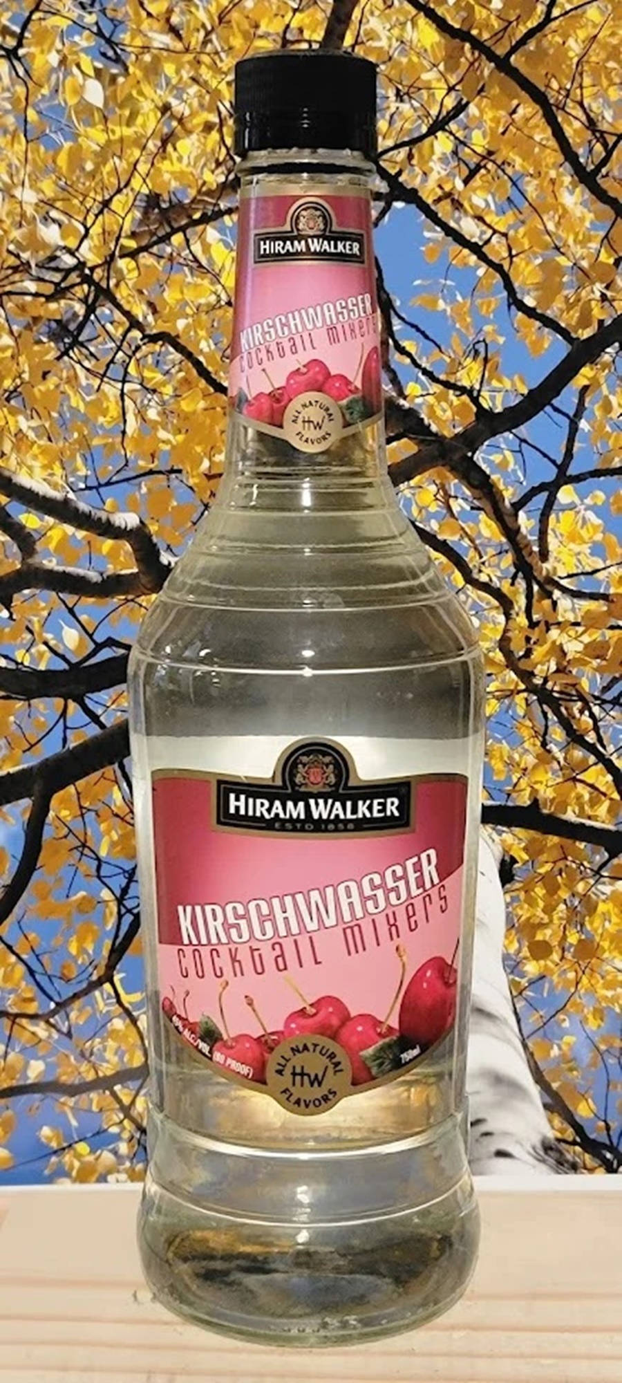 alcohol 桌布 壁紙 — 一瓶透明的Hiram Walker Kirschwasser雞尾酒調和劑，以金色秋葉和藍天為背景。
