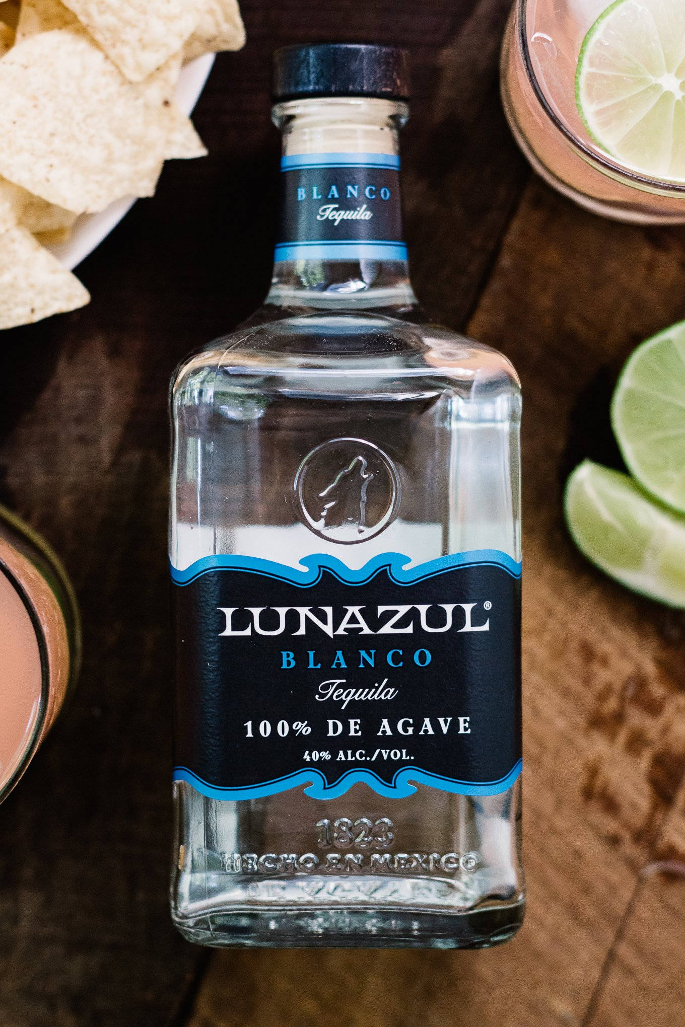 alcohol 桌布 壁紙 — 一瓶透明的Lunazul Blanco龍舌蘭酒放在深色木桌中央，周圍有萊姆角和玉米片。