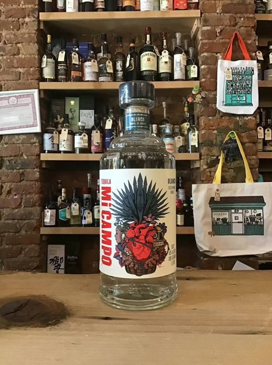 bottle 桌布 壁紙 — 一瓶透明的Mi Campo龍舌蘭酒，帶有鮮豔的紅色心形和龍舌蘭設計標籤，放在木製吧檯上，背景是磚牆和擺滿酒瓶的架子。