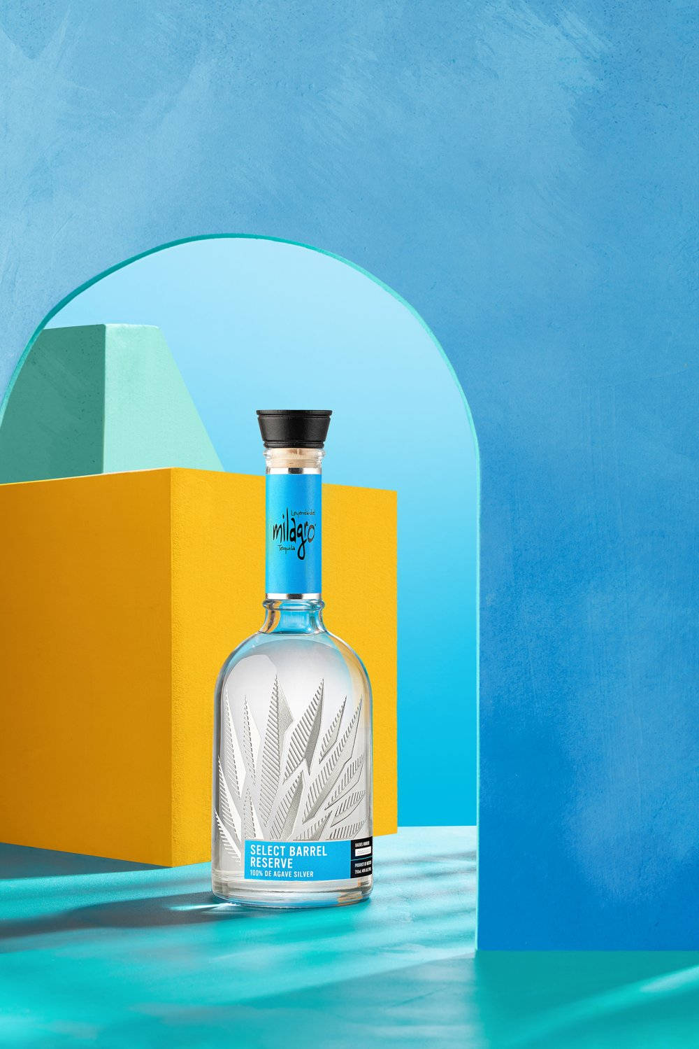 alcohol 桌布 壁紙 — 一瓶透明的Milagro Select Barrel Reserve龍舌蘭酒居中放置，背景為鮮豔的藍色，搭配大膽的黃色和藍綠色幾何形狀。