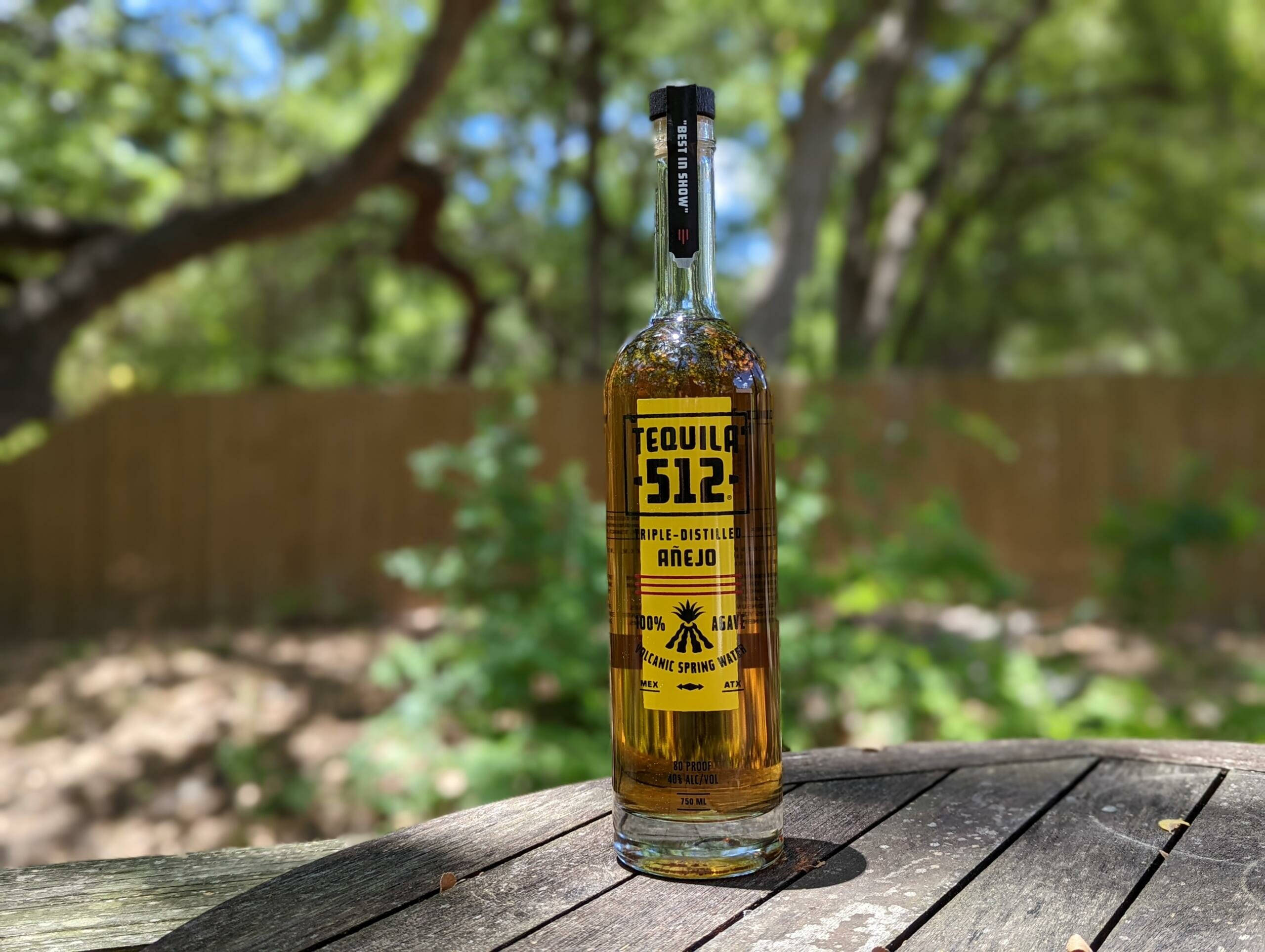 drinks 桌布 壁紙 — 一瓶透明的Tequila 512 Añejo放在戶外風化的木桌上，在明亮的陽光下，背景有模糊的綠樹。