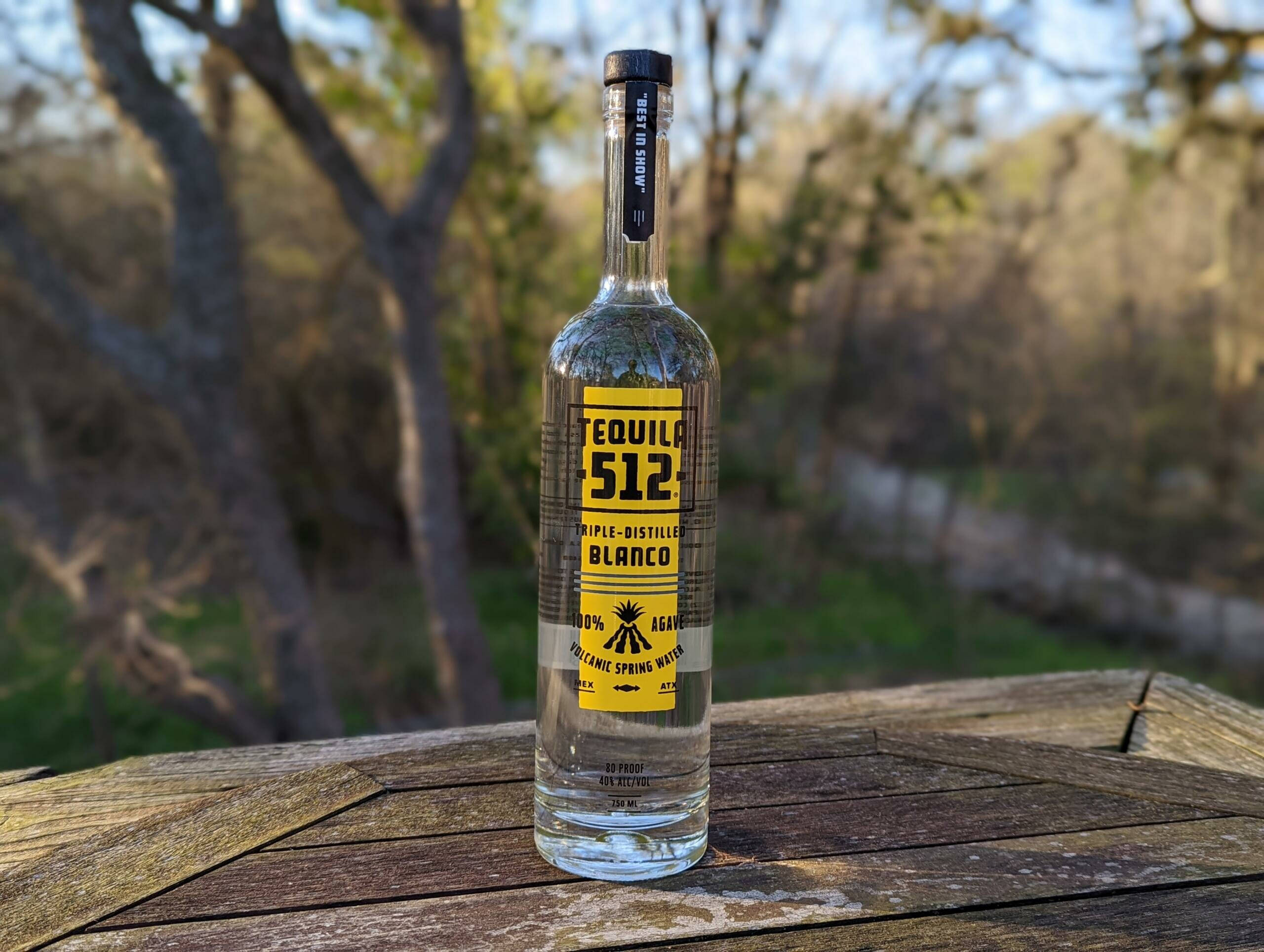 drinks 桌布 壁紙 — 一瓶透明的Tequila 512 Blanco酒瓶放在戶外一張風化的木桌上，背景有模糊的樹木和柔和陽光。