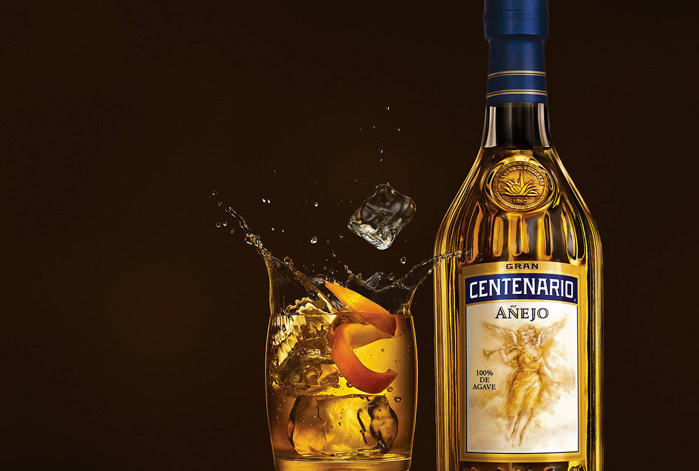 冰 桌布 壁紙 — 一瓶金色的Centenario Añejo龍舌蘭酒放在玻璃杯旁，冰塊濺入琥珀色液體中，帶有柑橘風味。