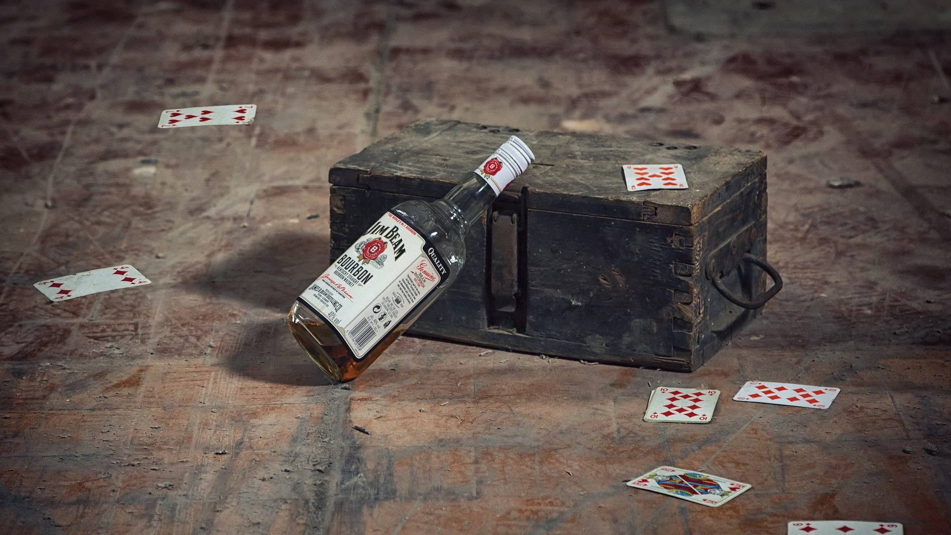 Poker 桌布 壁紙 — 一瓶風化的波本威士忌靠在一個質樸的木箱上，在磨損的地板上，周圍散落著撲克牌。