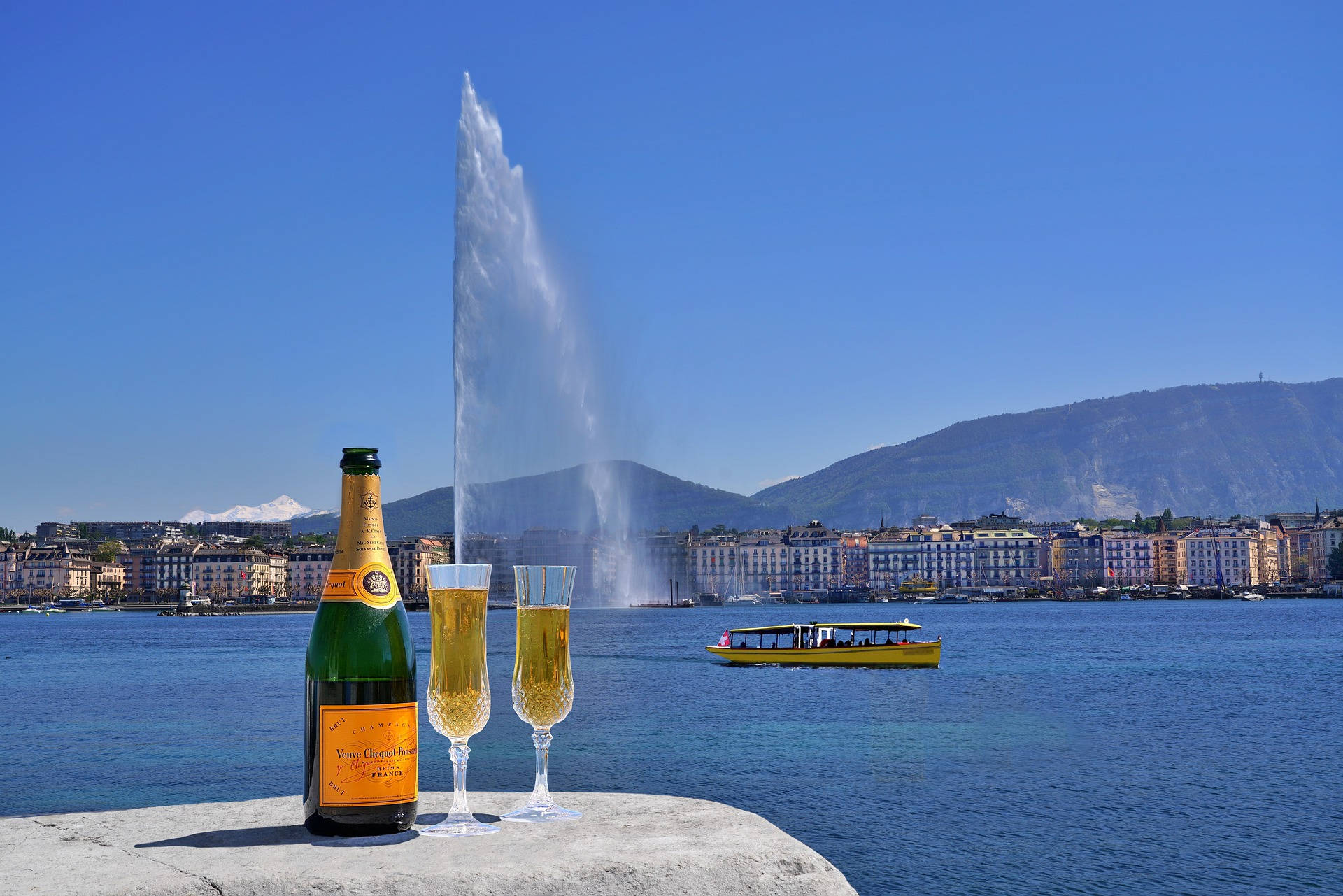 Champagne 桌布 壁紙 — 一瓶香檳和兩個酒杯放在石檯上，俯瞰日內瓦湖，標誌性的 Jet d'Eau 噴泉在清澈的藍天下高高噴射。