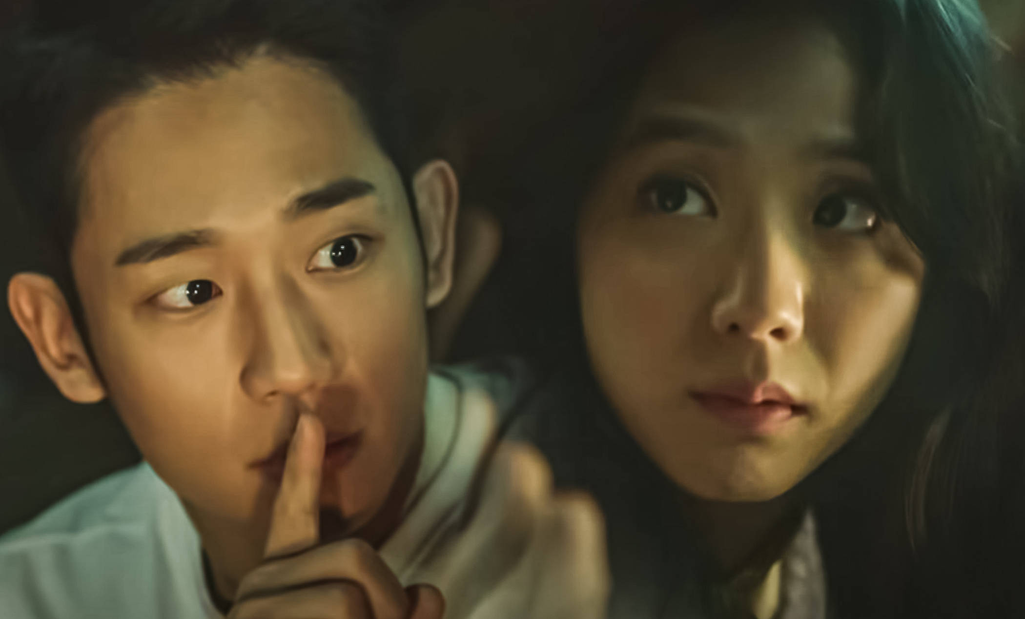 drama 桌布 壁紙 — 一男一女在昏暗燈光下分享一個安靜而緊張的時刻，男子將手指放在嘴唇上。