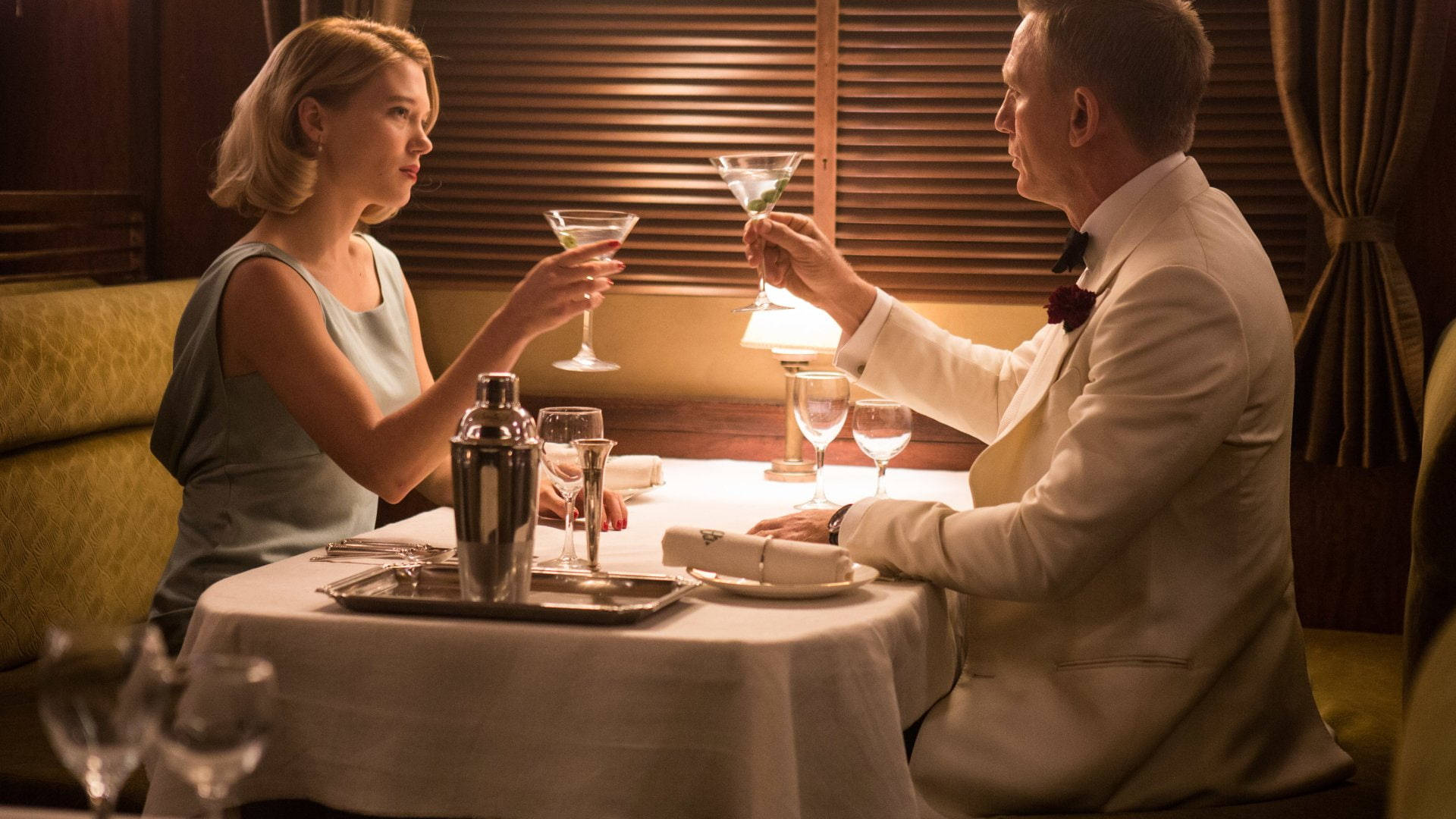 Daniel Craig 桌布 壁紙 — 一男一女在溫暖燈光的高級餐廳卡座中碰杯馬丁尼酒杯，配有木百葉窗和黃色軟墊。
