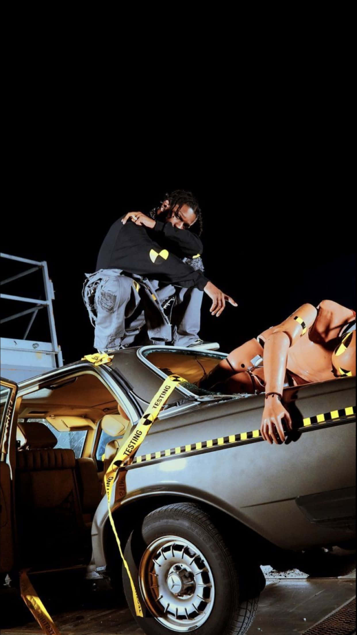 ASAP Rocky 桌布 壁紙 — 一男一女在黑暗夜空下，以戲劇性的姿勢站在一輛被黃色警示膠帶包裹的廢車上。
