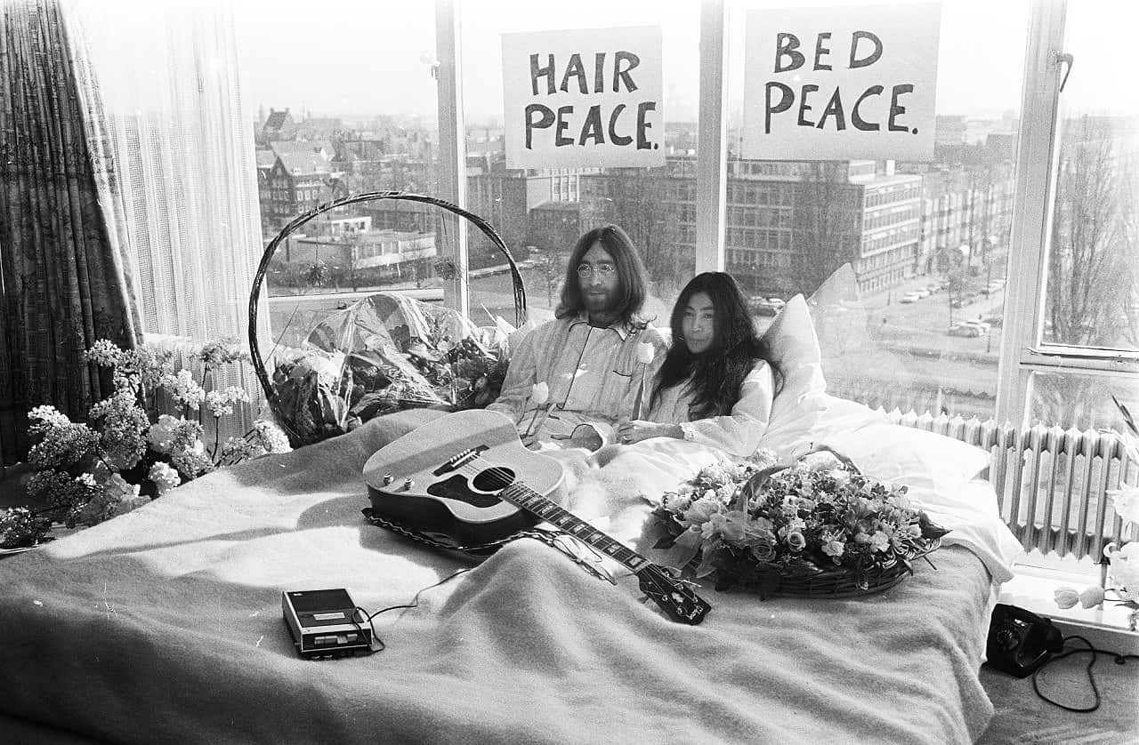 和平 桌布 壁紙 — 一男一女坐在床上，拿著吉他和鮮花，舉著寫有「HAIR PEACE」和「BED PEACE」的牌子，房間陽光充足，可看到城市景觀。