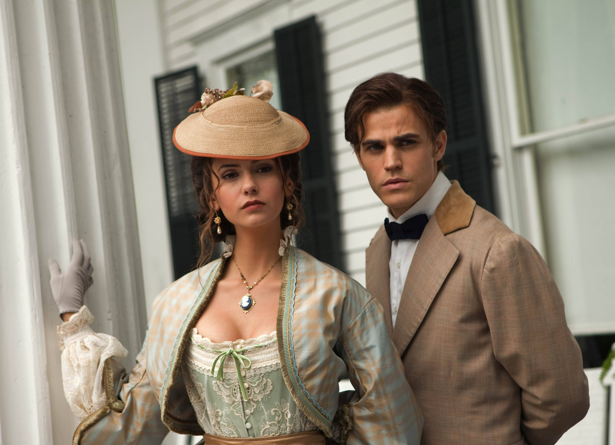 The Vampire Diaries 桌布 壁紙 — 一男一女穿著復古服裝，莊嚴地站在白色牆板和黑色百葉窗的門廊上。