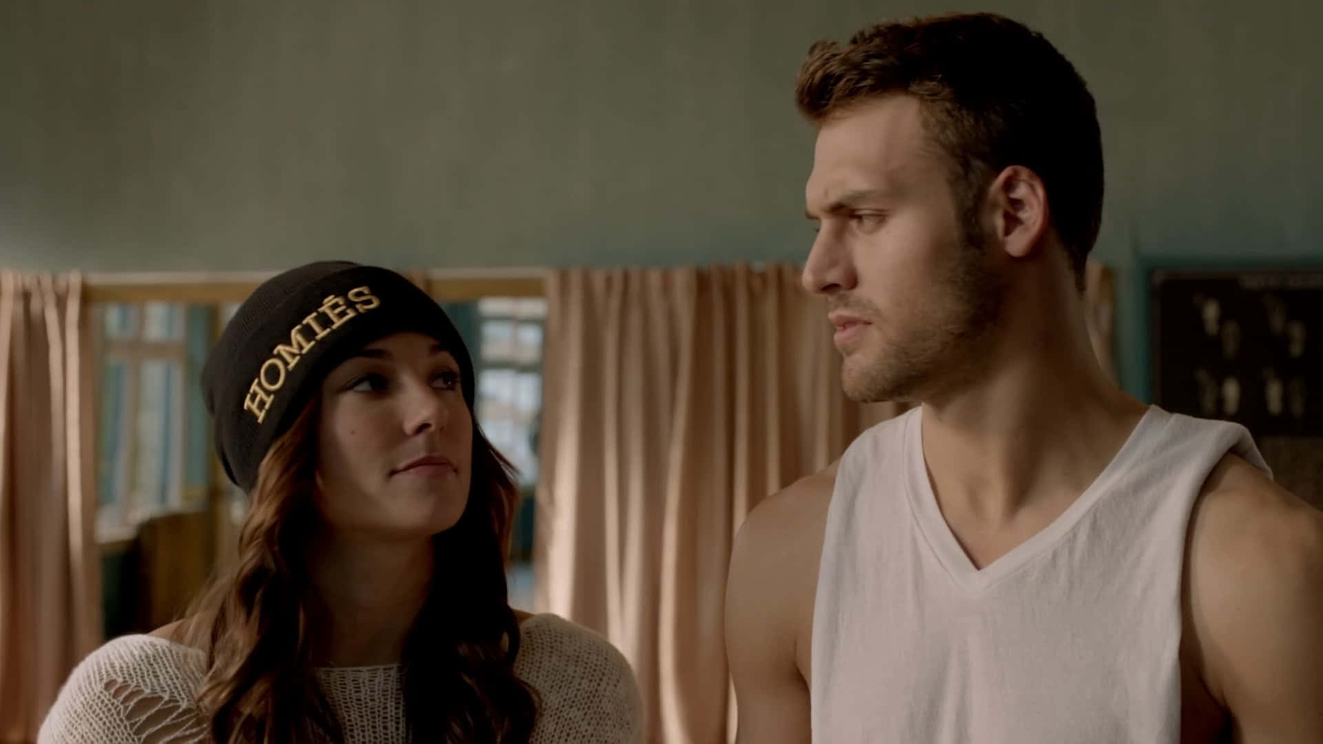 Briana Evigan 桌布 壁紙 — 一男一女站在燈光柔和的房間裡，女子戴著「HOMIES」毛帽，彼此靜靜地凝視對方。