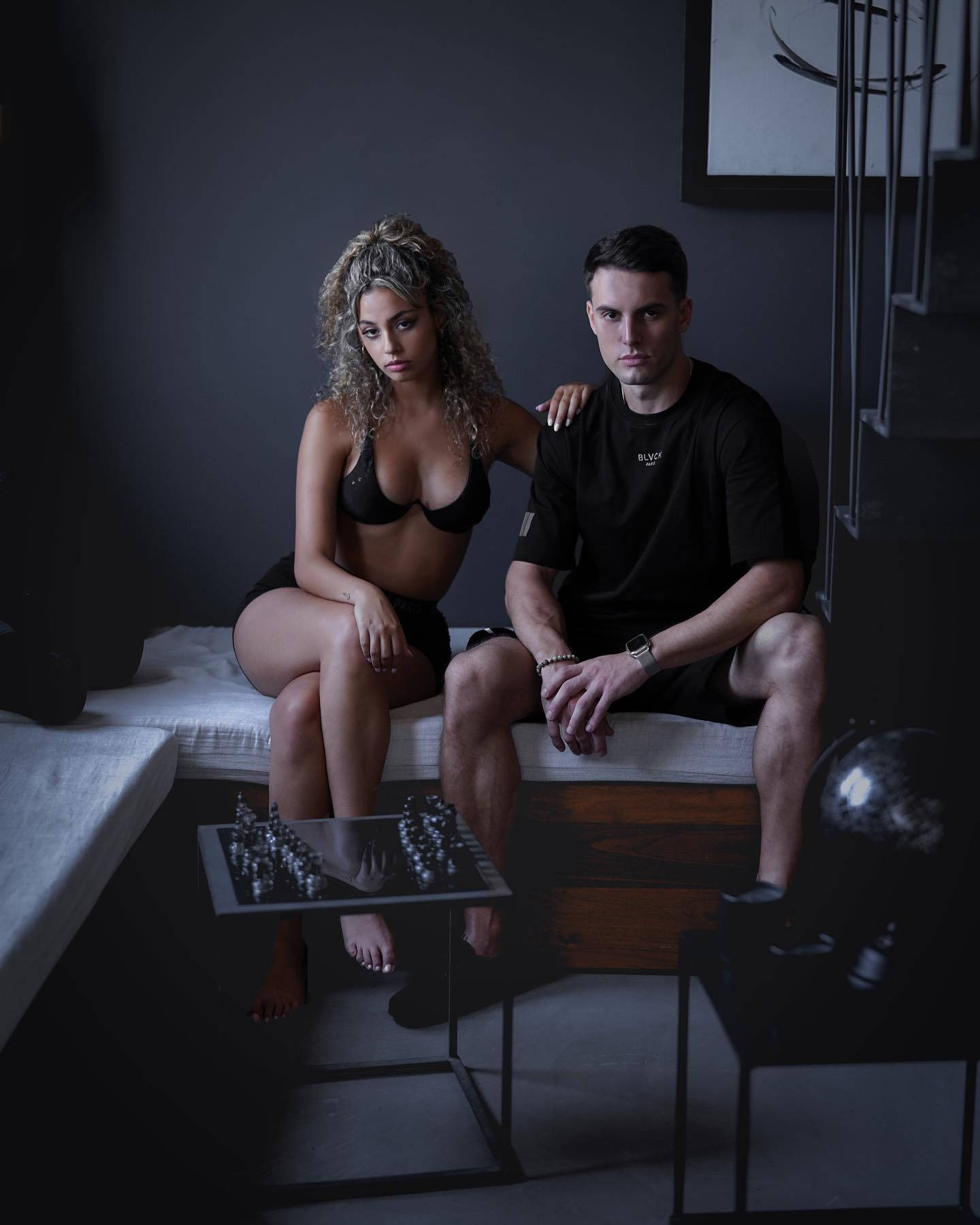 Blvck Paris 桌布 壁紙 — 一男一女親密地坐在光線昏暗的現代房間中的白色長椅上，玻璃桌上放著西洋棋棋子。