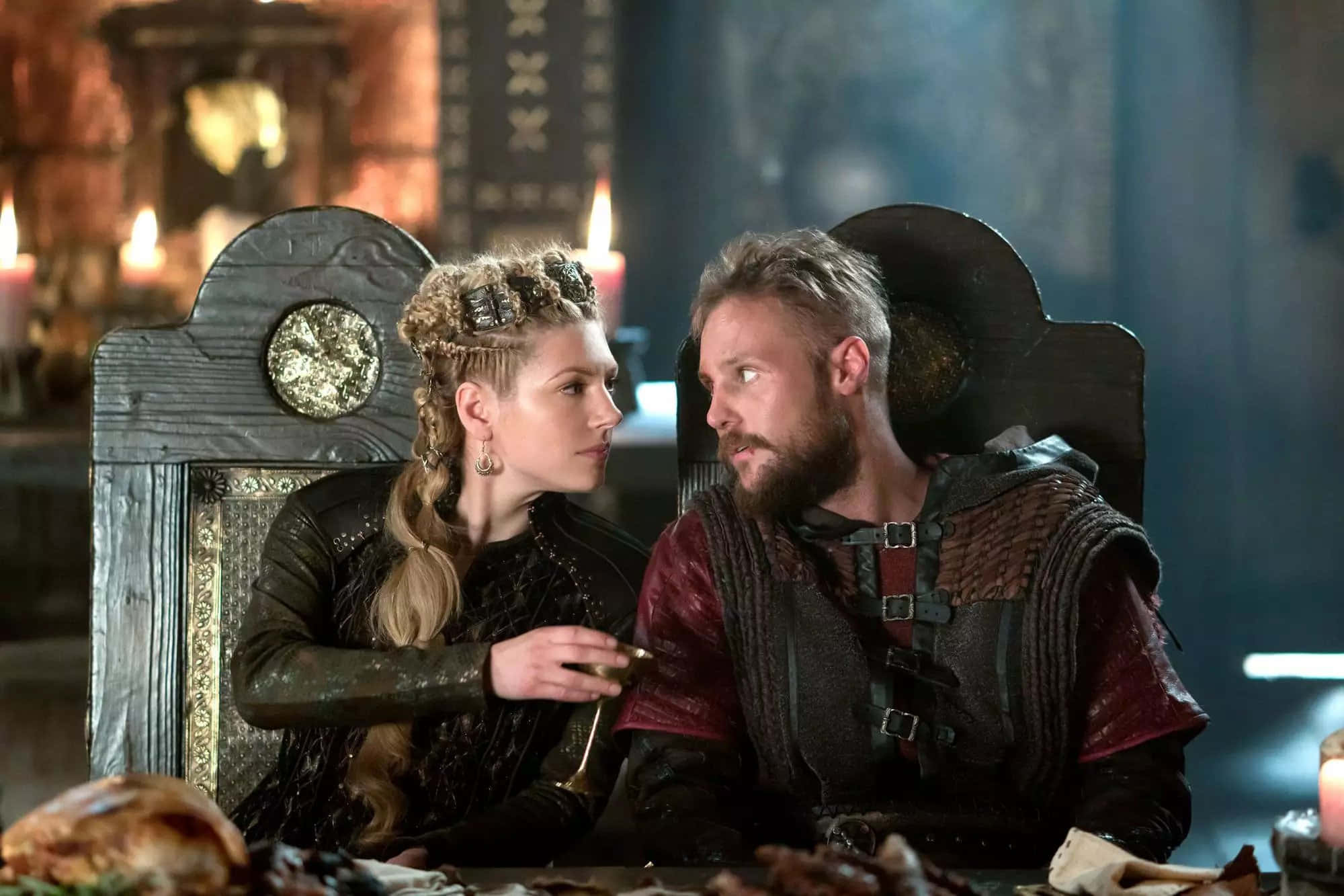 Vikings 桌布 壁紙 — 一男一女身穿中世紀盔甲，在燭光搖曳的寶座大廳中隔桌而坐，目光交織，氣氛緊張。