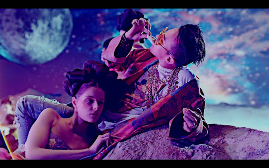 G-dragon 桌布 壁紙 — 一男一女躺臥在岩石上，下方是宇宙天空，有巨大的月亮和星雲，閃爍著鮮豔的紫色和藍色。
