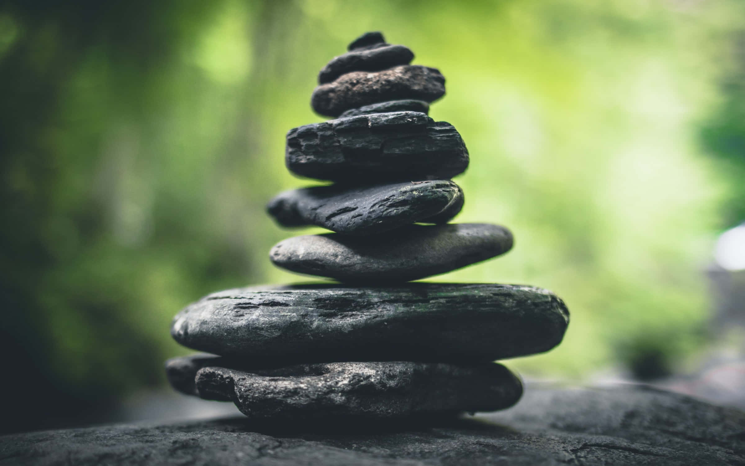 禪 桌布 壁紙 — 一疊光滑的深色石頭優雅地平衡在柔和的綠色森林背景上。