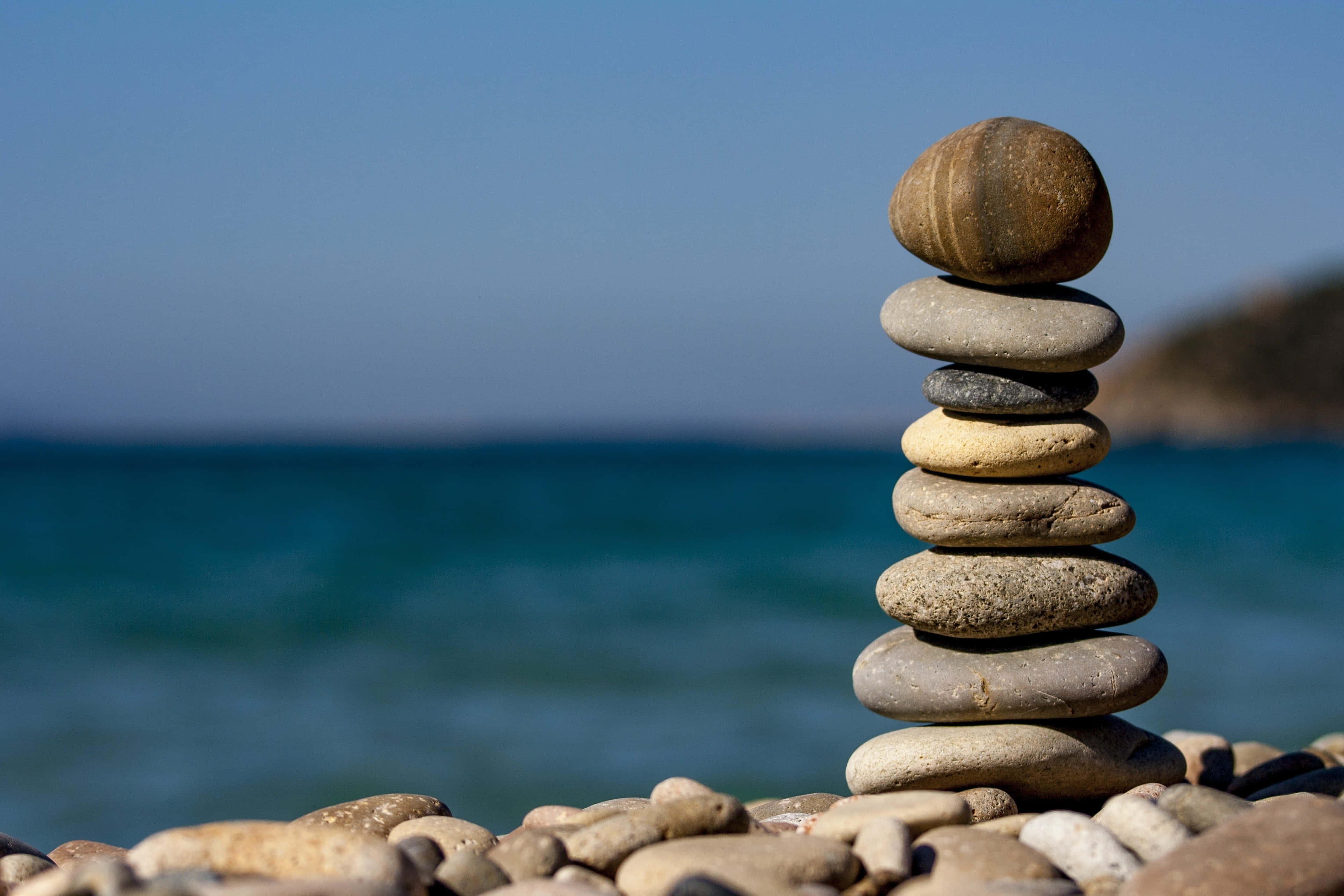 放鬆身心，享受這片寧靜背景。 壁紙 — 一疊光滑的石頭在鵝卵石海灘上平衡，背景是柔焦的碧綠海洋和清澈的藍天。