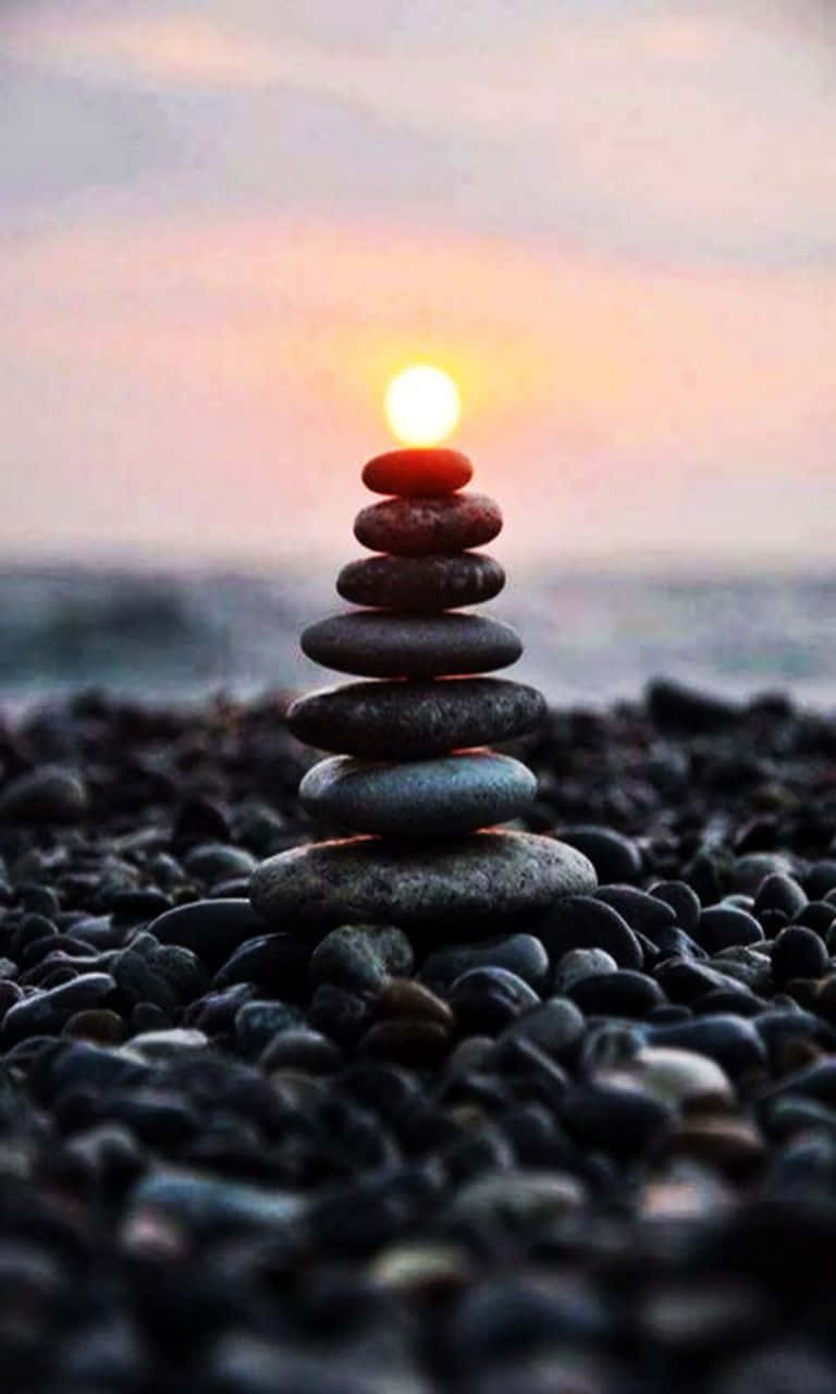 禪 桌布 壁紙 — 一疊光滑的石頭在鵝卵石海灘上平靜地保持平衡，同時一個發光的球體從頂部的石頭升起，映襯著柔和的日落天空。