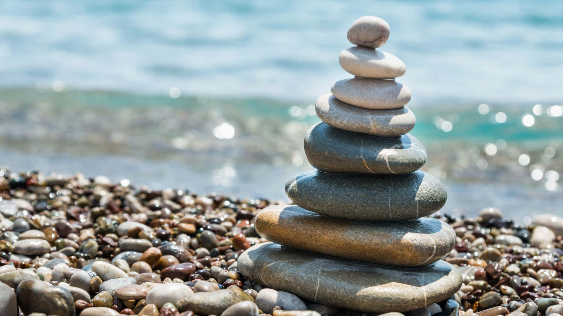 Relaxing Desktop 桌布 壁紙 — 一疊完美平衡的光滑石頭從陽光照耀的卵石海灘上聳立，背景是溫柔的海浪。