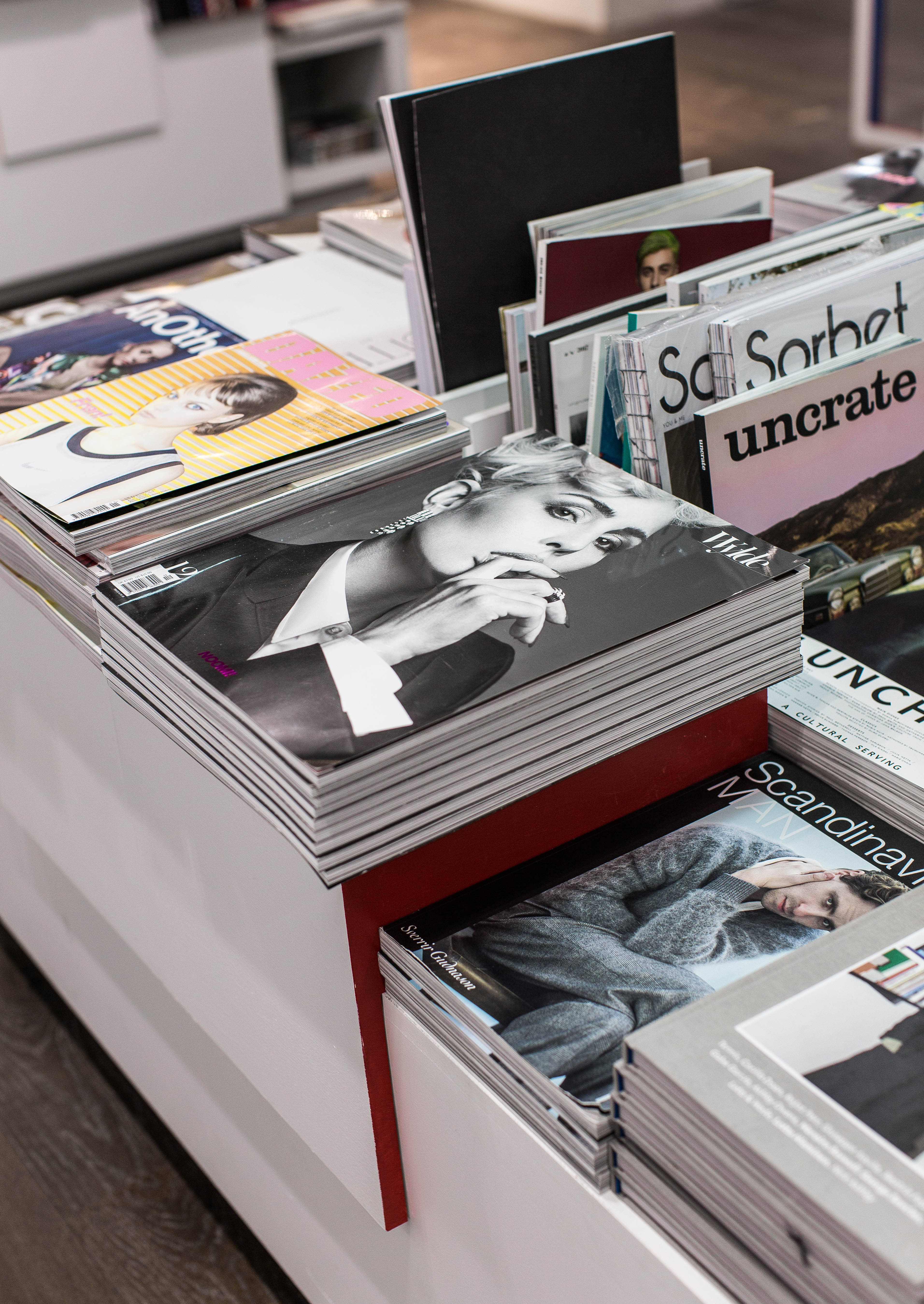 Magazine 桌布 壁紙 — 一疊疊光面雜誌，封面大膽，放在現代白色架子上，位於光線充足的零售空間。