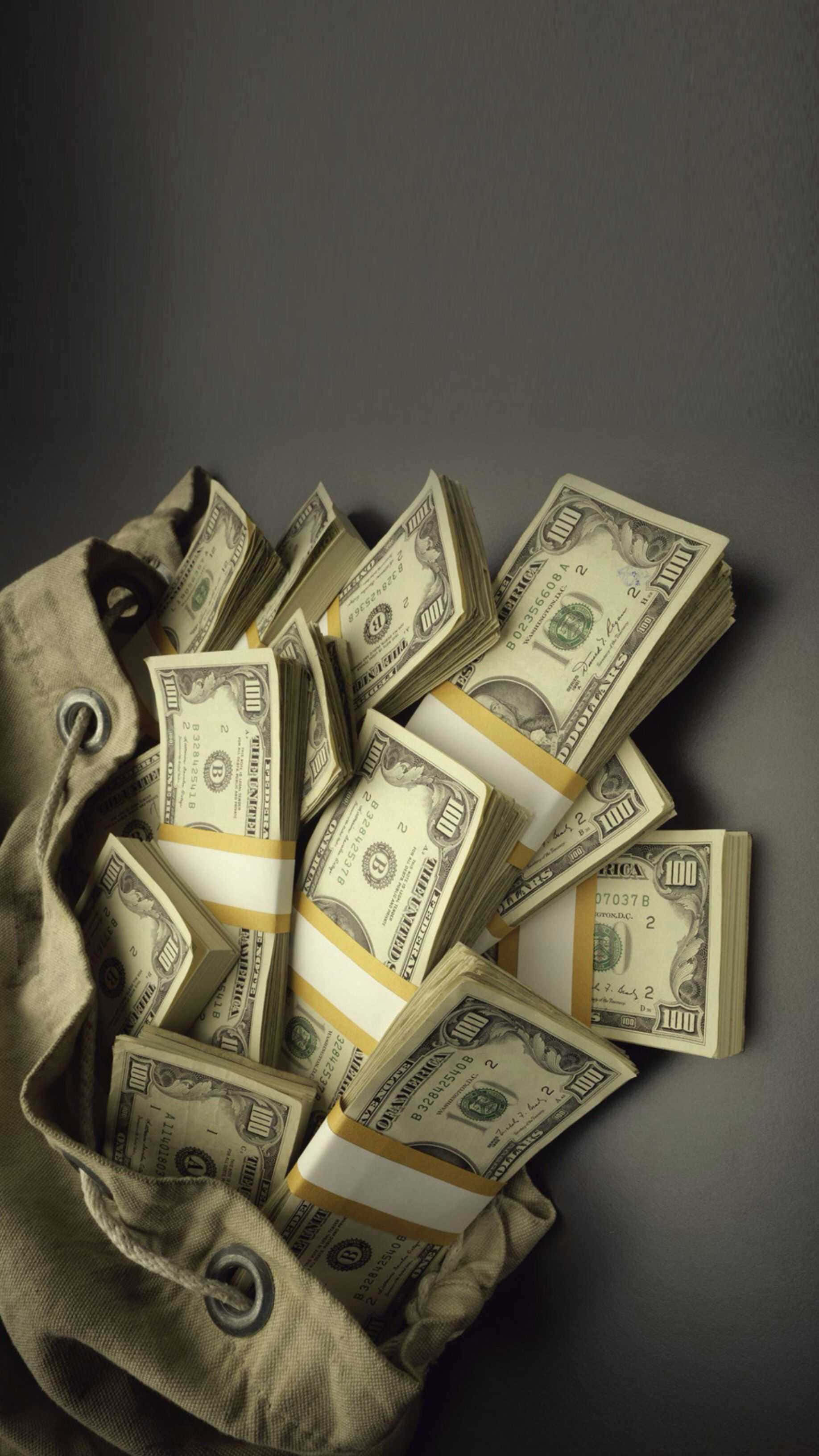 金錢 桌布 壁紙 — 一疊疊百元美鈔從米色帆布袋中溢出，背景為深灰色。