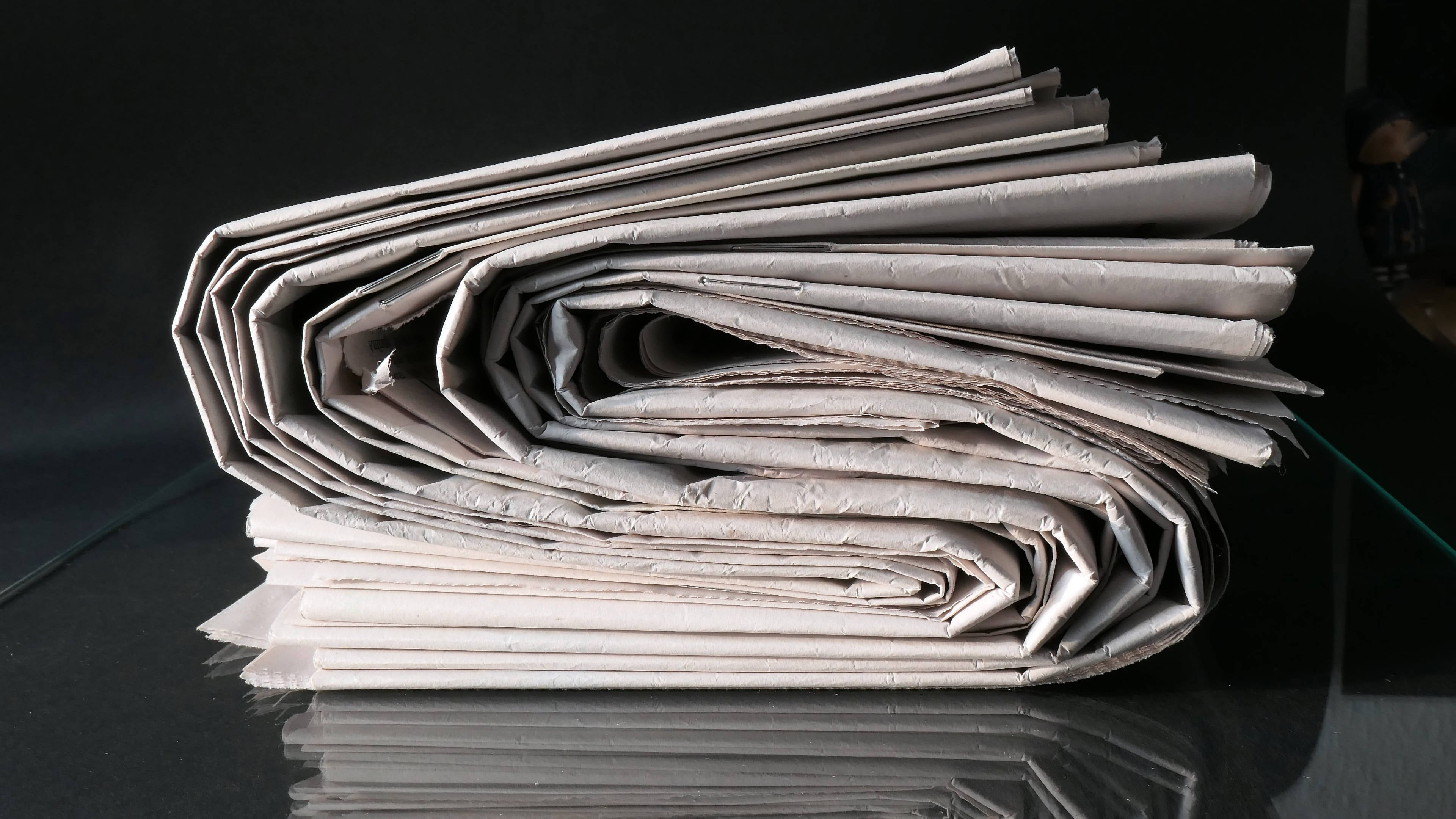Newspaper 桌布 壁紙 — 一疊緊密捲起的報紙頁面放在光滑的黑色表面上，帶有清晰的反射。