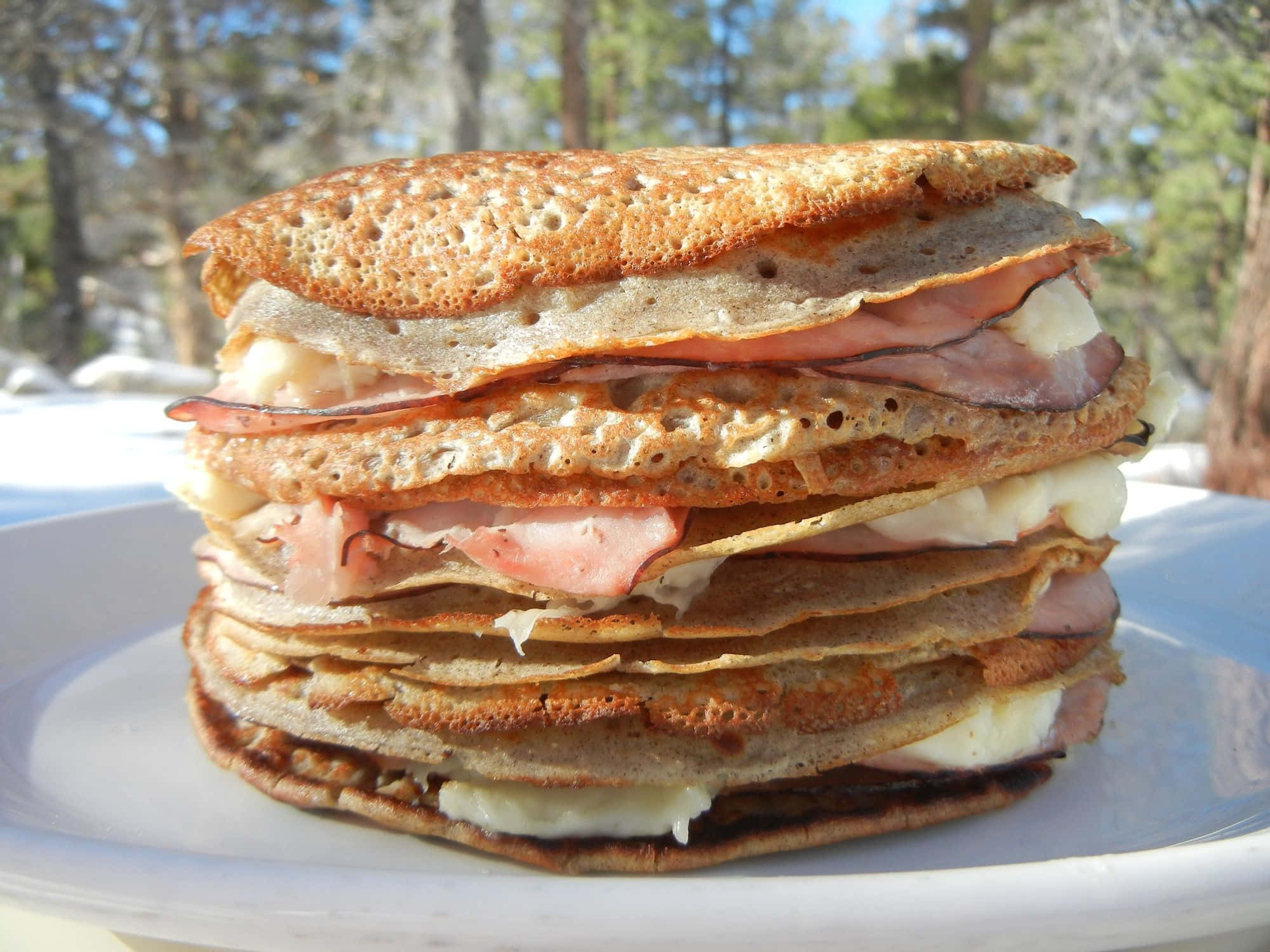 Black Forest Ham 桌布 壁紙 — 一疊高高的金黃色可麗餅，內餡是融化的起司和火腿，放在白色盤子上，在戶外明亮的陽光下。